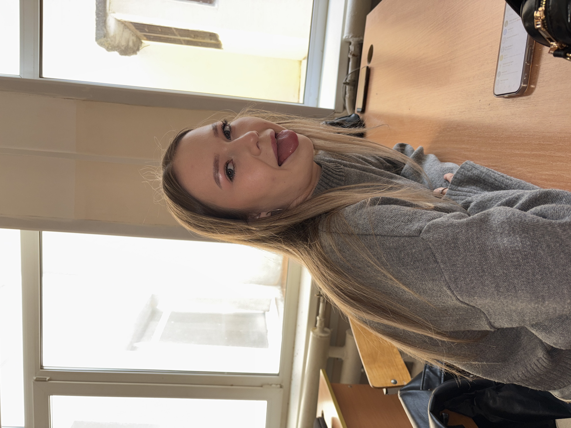 A young woman with long blonde hair, wearing a gray sweater, is sitting at a wooden desk next to a window in what appears to be a classroom. She is sticking her tongue out. A cell phone and a black bag are visible on the desk. The location appears to be a building in Chaikovsky, Russia, as suggested by metadata. A chair can also be seen in the background.