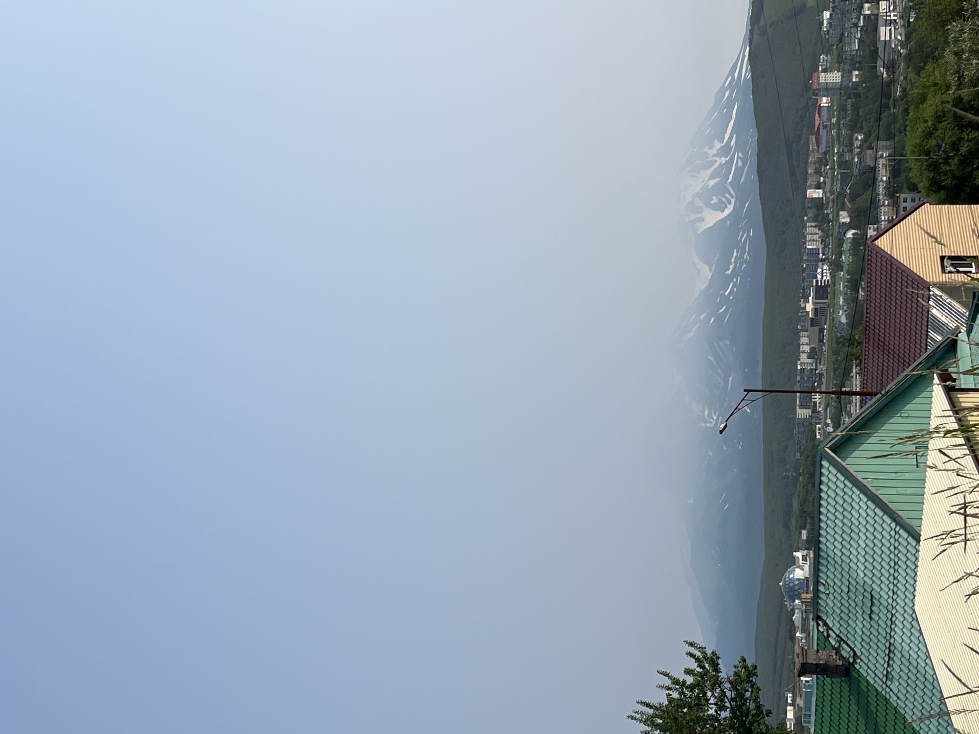 The image shows a panoramic view of the city of Petropavlovsk-Kamchatsky, Russia, with a mountain range in the background. In the foreground, there are several houses with colorful roofs, and a tree on the left side. The middle ground features a collection of buildings, including a distinctive dome-shaped structure and a street lamp. The background is dominated by a large, snow-capped mountain range partially obscured by clouds. The sky is clear and light blue.