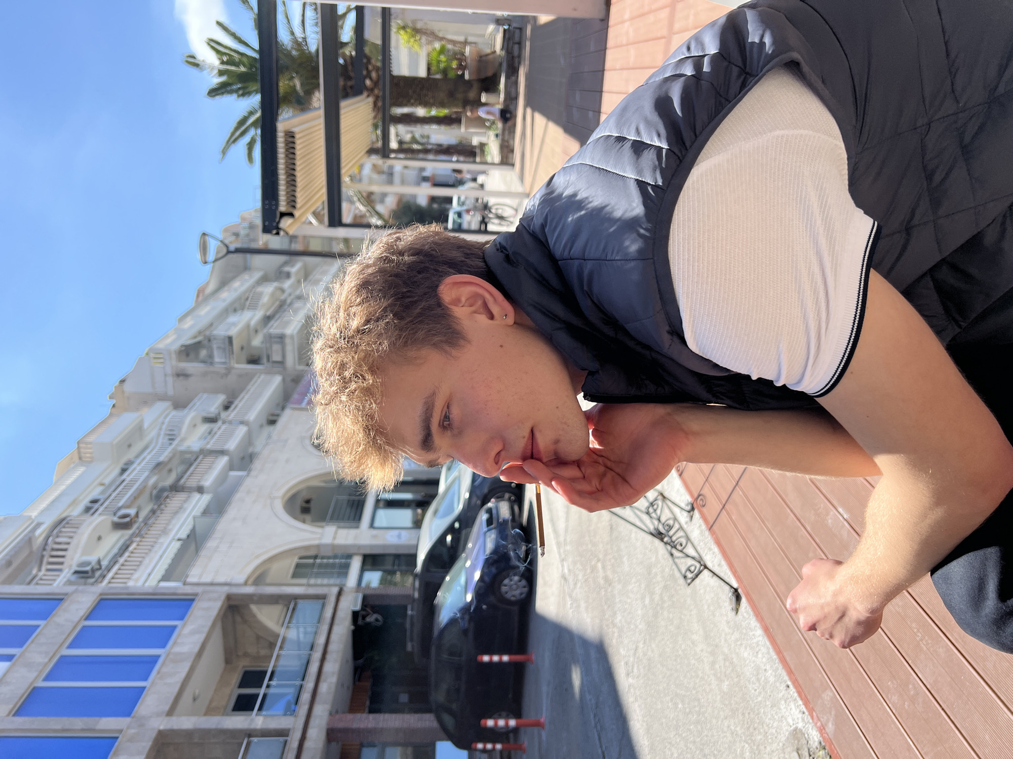 A young man is pictured in Budva, Montenegro, with a cigarette between his fingers. He is wearing a dark-blue quilted vest over a white short-sleeved shirt with dark trim at the sleeve. The building across the street, visible behind a black car, has a modern architectural style. A palm tree and a covered outdoor seating area are also visible, suggesting a warm climate. The ground has wood planks, indicating an outdoor deck or patio-like space. The sky is clear and blue.