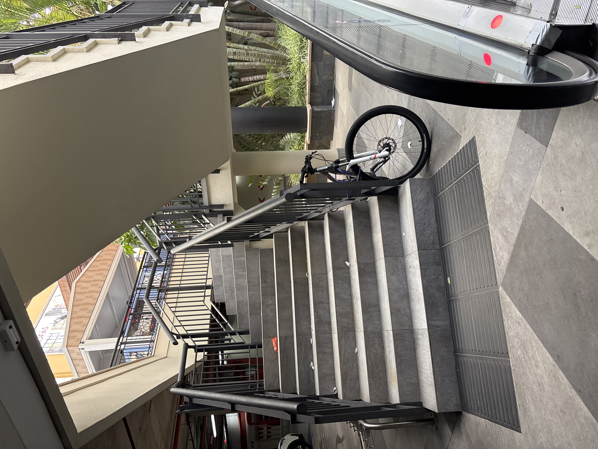 The image shows a public outdoor stairway with a bicycle parked near its base, next to an escalator. The stairway is constructed with gray stone tiles and has black metal railings on both sides. The bicycle is a mountain bike with a black frame, white front forks, and black wheels. It is leaning against the railing. In the background, there are palm trees and other tropical-looking plants, suggesting a warm climate. The ground is paved with gray tiles. There is a red plastic bag on one of the steps. The scene appears to be in a commercial or recreational area.