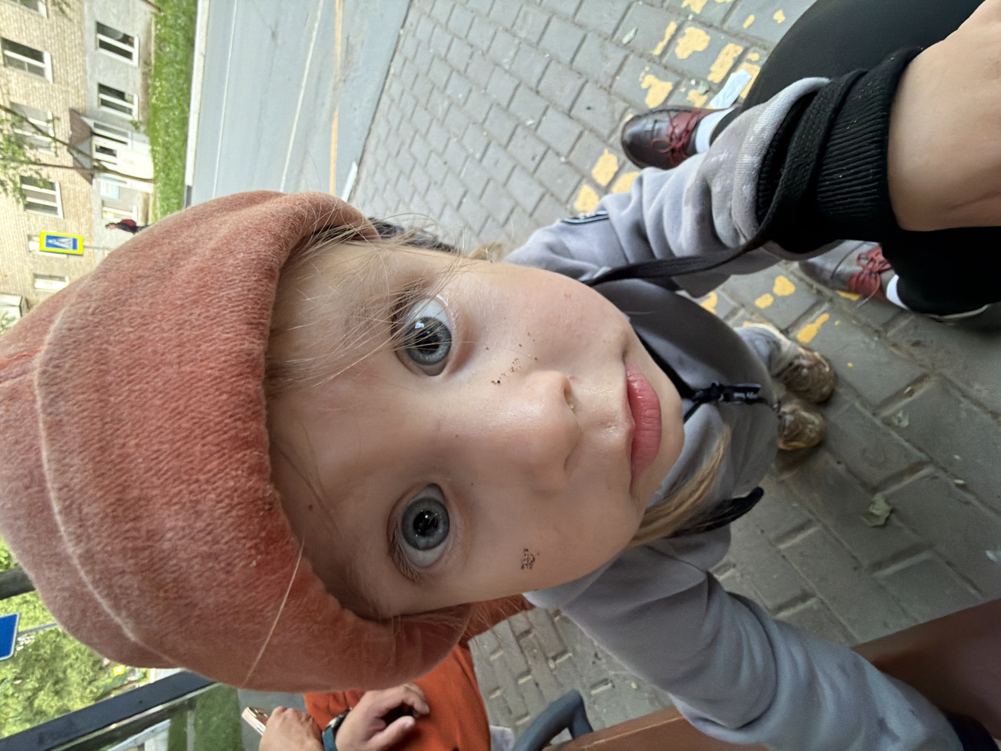 A close-up, high-angle outdoor shot captures a young child, likely a girl, looking directly up at the camera. She wears a textured, salmon-colored hat or bonnet and a light grey hooded sweatshirt. Her striking blue eyes are wide open, and her lips are slightly parted, revealing a soft pink color. Scattered specks of dirt are visible on her nose and right cheek, suggesting she has been playing outdoors.
The shot is taken from a very low perspective, possibly by an adult holding the camera or the child looking up from a lower position. Part of an adult's body is visible on the right, including a black pant leg, a grey shoe with dark red laces, and a sleeve with a black cuff. Another foot is visible further back, wearing a grey shoe.
The background shows an urban or suburban setting. Behind the child, a paved area with grey block-style tiles and yellow markings stretches out. Beyond that is a road with a grey surface and faint yellow lines. On the far left, multi-story residential buildings with multiple windows are visible, partially obscured by green trees. A blue and yellow road sign is also faintly discernible in the distance. The lighting suggests it is daytime, possibly overcast, creating soft illumination. The overall scene conveys a casual, candid moment outdoors in Chaikovsky, Russia. No legible text is visible in the image.