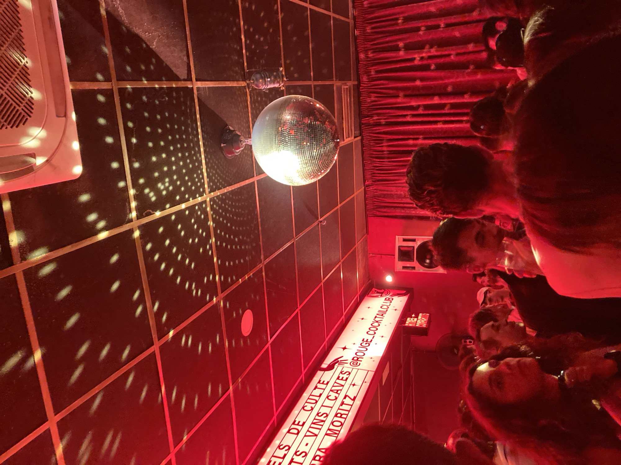 The image shows the interior of a dimly lit space, seemingly a nightclub or cocktail bar, possibly located in Barcelona, Spain, indicated by the sign referencing @ROUGE_COCKTAILCLUB, along with "Vins Caves", "Moritz". The predominant light source is a disco ball hanging from the ceiling, which reflects light onto the dark tiled floor. The floor is a grid of dark tiles separated by thin, light-colored lines, with small, scattered spots of light reflected from the disco ball. Near the upper-left of the frame, there is an air conditioning unit. On the right side, there are a group of people, primarily seen from the chest up, with their faces visible. The room is illuminated by red light, with some red curtains in the background.