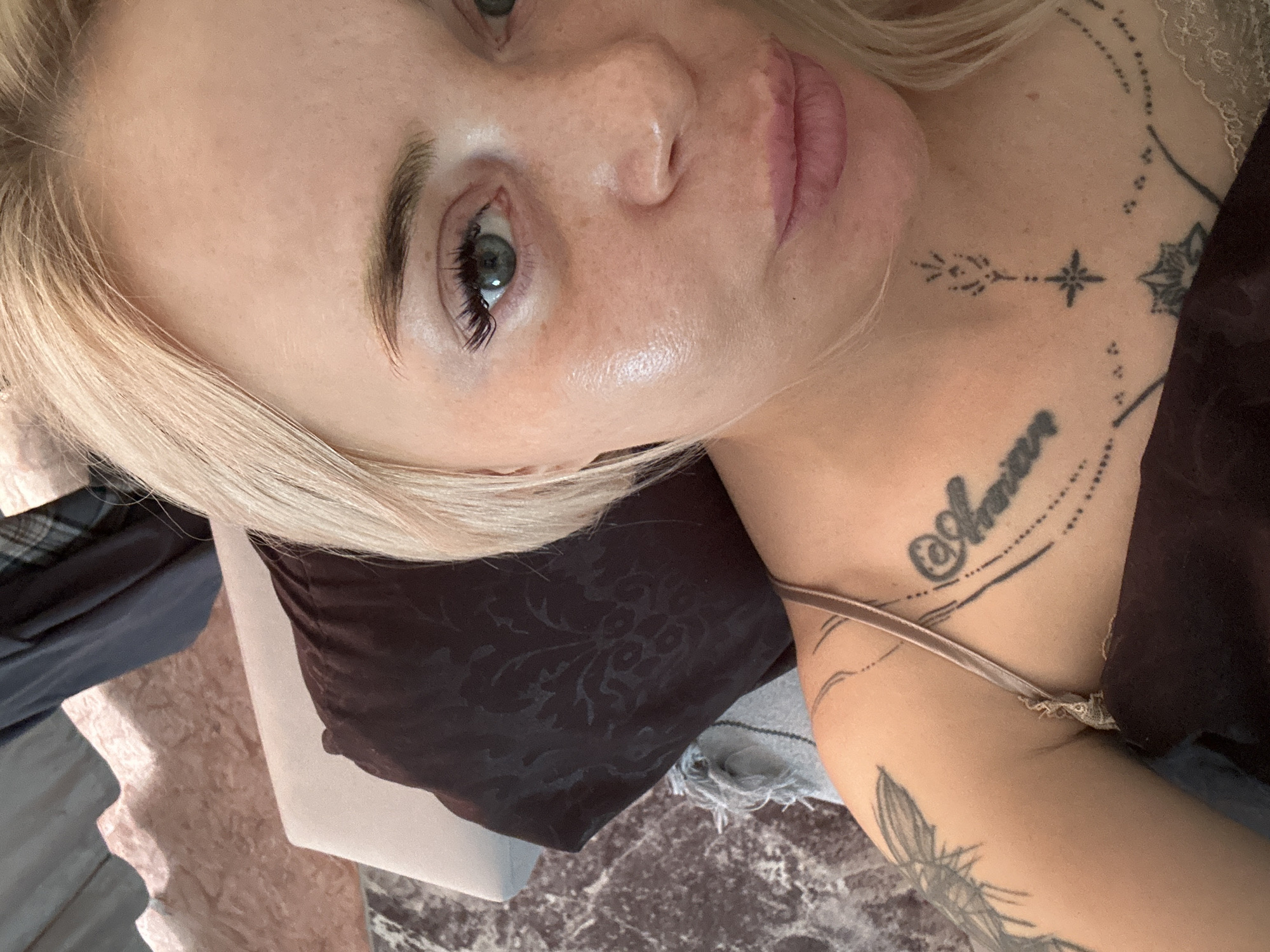 A fair-skinned individual with blonde hair, estimated to be female, is captured in a close-up, high-angle selfie. The subject's gaze is directed towards the camera, with the left eye, which is green or blue, fully visible and framed by prominent eyelashes. The lips are full, pink, and slightly pursed. The skin exhibits a visible sheen on the left cheek and forehead, along with scattered freckles. Multiple tattoos are present: a large insect design on the upper left arm/shoulder, the word "Amor" in a script font on the upper right chest, a central geometric star-like design on the collarbone, and dotted line patterns forming an arc with a partial lotus design below it. The subject is wearing a brown or bronze colored spaghetti-strap garment, possibly satin, featuring a subtle lace trim on the right side. The background indicates an indoor environment, featuring a dark brown pillow with a floral damask pattern, a dark mottled surface resembling marble, and a textured light brown wall or furniture with a wavy edge, alongside dark fabric and a plaid pattern in the upper right. The location is in Unknown, Unknown.