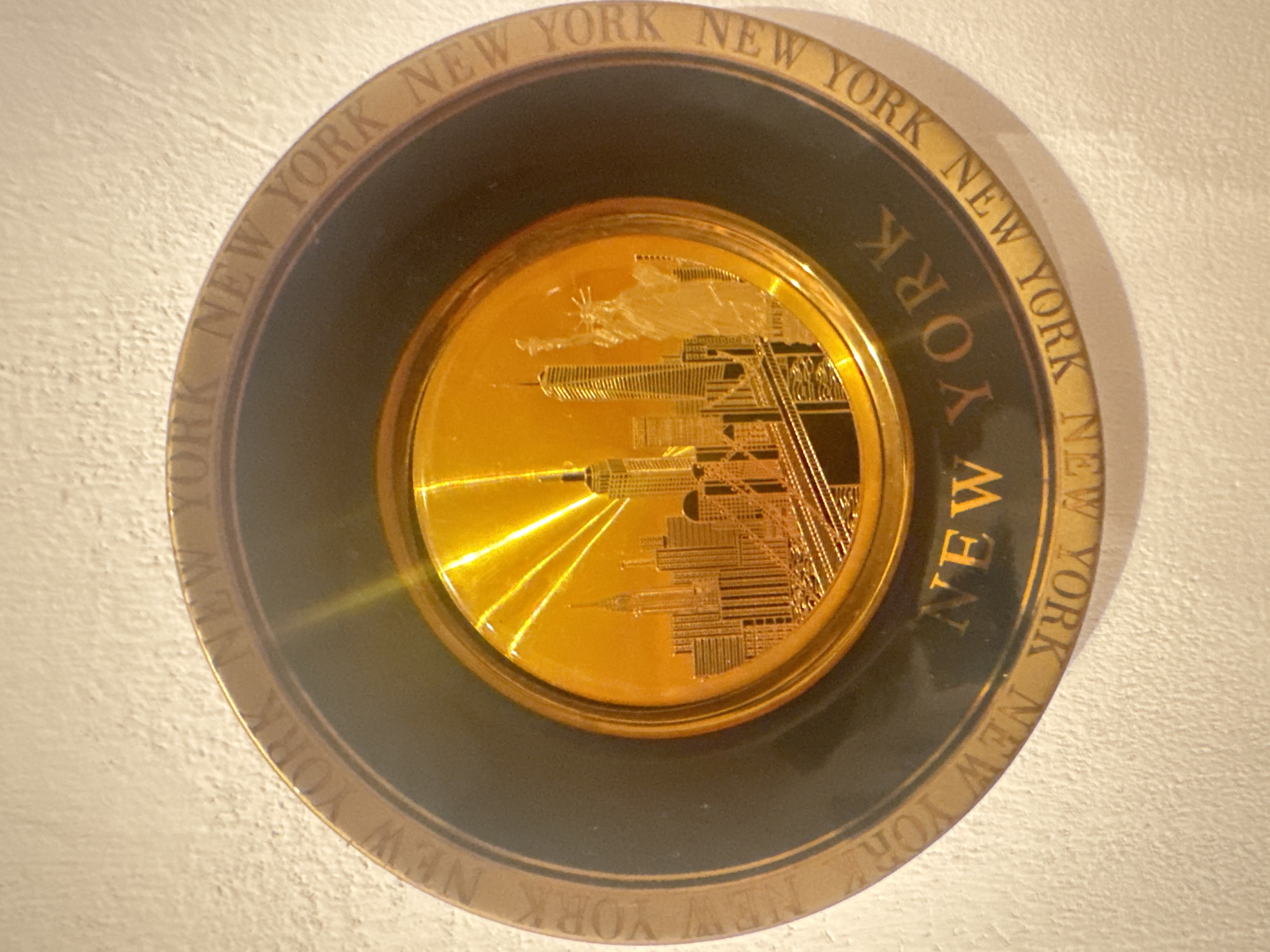 A circular, gold-colored plate with a black border hangs on a textured white wall. The plate features a detailed embossed image of the New York City skyline, including skyscrapers and the Statue of Liberty, set against a background with radiating lines suggesting sunlight. The words "NEW YORK" are embossed around the outer edge of the plate in a circular pattern. The plate is illuminated by a light source that creates glare and reflections on its surface.