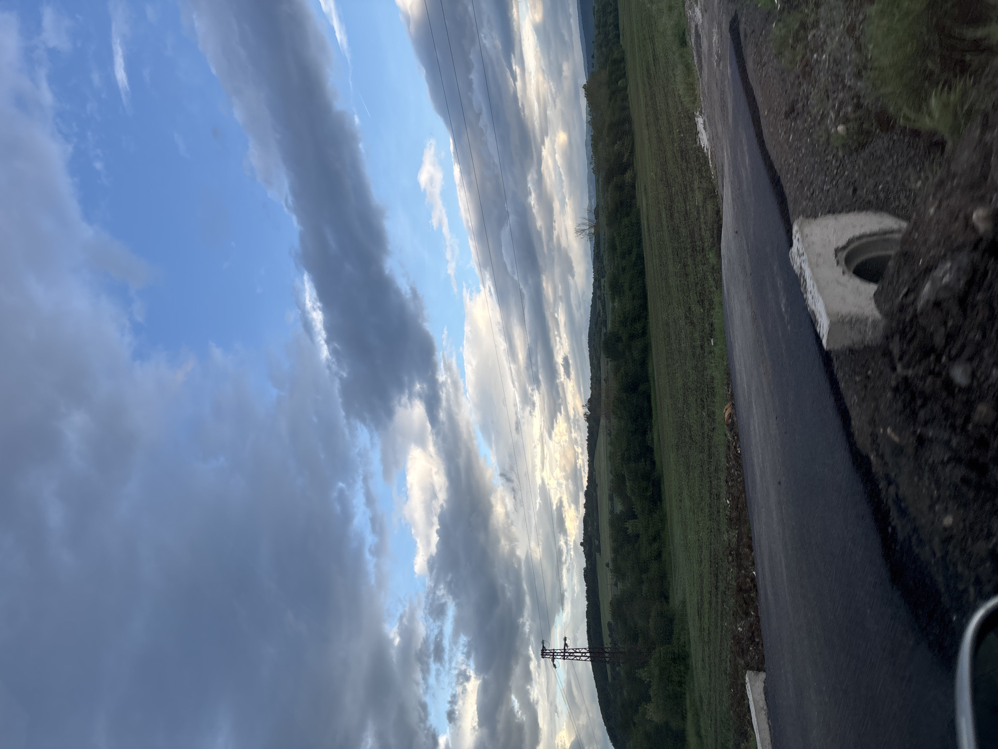 The image depicts a rural scene in Belopoptsi, Bulgaria. A newly paved asphalt road runs along the right side of the frame. Adjacent to the road, there is a concrete structure, possibly a drain or manhole. To the left of the road stretches a green field bordered by a line of trees or forest. The sky occupies the majority of the image and is filled with a mix of blue sky and gray clouds. Overhead power lines are visible crossing the sky and a utility pole.