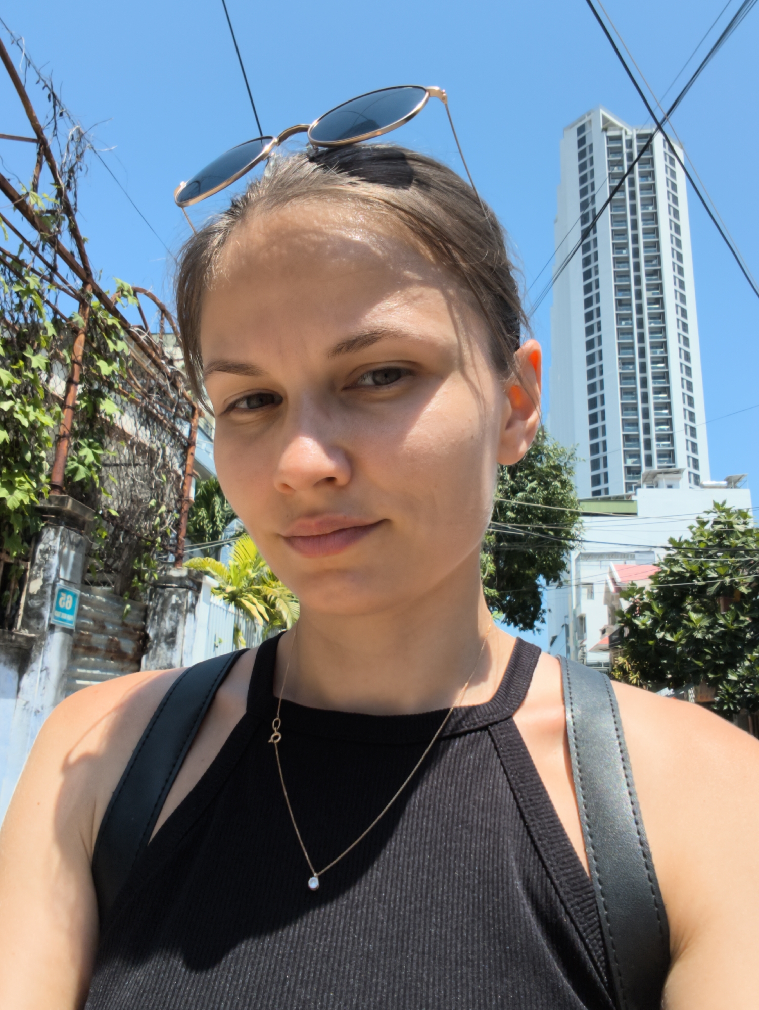 A young woman is taking a selfie outdoors. She is wearing a black sleeveless top and a delicate gold necklace with a small pendant. Sunglasses are perched on top of her head. Her hair is pulled back, and she is looking at the camera with a slight smile. In the background, a tall, modern white building with many windows dominates the skyline, indicating an urban setting. To the left, there is a weathered wall covered in vines and a blue sign with white lettering. Lush green trees are visible on the right side of the frame. The sky is clear and blue, suggesting a sunny day. This scene is located in the city of Nha Trang Ward, Vietnam.