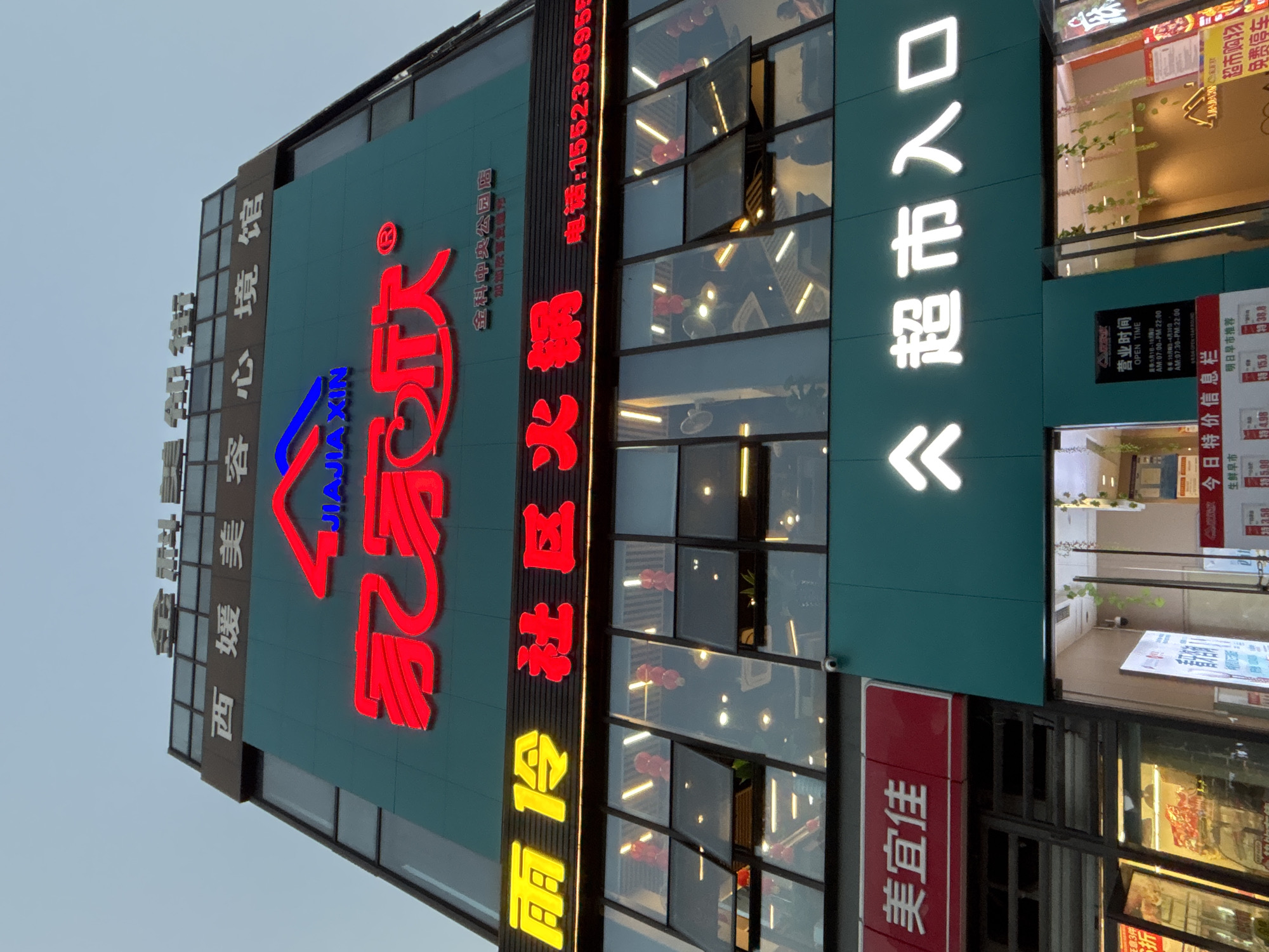 The image depicts the exterior of a building in the city of Jiangjin, China, identified by signage indicating “西媛美容心境馆” (Xi Yuan Beauty Mind Museum), “Jia Jia Xin” (a business or brand name) and “加加欣” (Jia Jia Xin), and “金科中央公园店”(Jinko Central Park Store). The building also houses a restaurant specializing in “雨令社区火锅” (Yuling Community Hot Pot) The telephone number associated with the hot pot restaurant is 1552398955. The presence of a "美宜佳" (Mei Yi Jia) convenience store and a sign indicating “超市入口” (Supermarket Entrance) suggests the building functions as a commercial space. Opening hours are listed. Various advertisement posters appear. The time of day appears to be early evening.