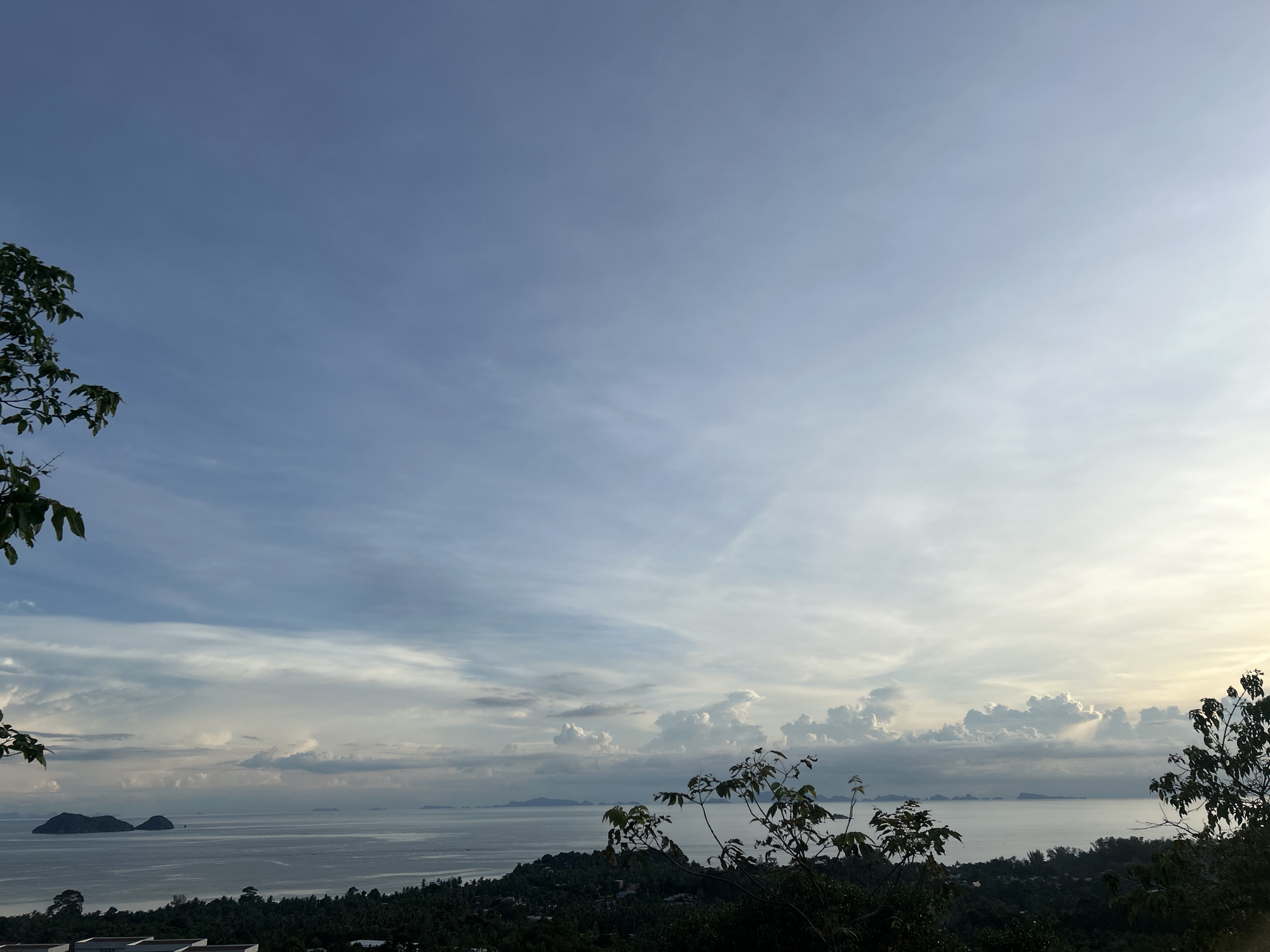 This media file captures a scenic view of a coastal landscape, likely in Koh Phangan, Thailand. The scene unfolds under a vast sky, transitioning from a clear, pale blue in the upper portion to more dramatic cloud formations nearer the horizon. Wispy cirrus clouds are interspersed with larger, puffy cumulus clouds, some of which appear to be illuminated by the sun from behind, creating a soft, diffused light.
The foreground is dominated by lush, dark green foliage, with branches and leaves of trees reaching into the frame from the left and right sides, framing the broader vista. This vegetation suggests a tropical or subtropical environment.
Below the sky and foliage lies a wide expanse of calm, greyish-blue water, representing the sea. In the distance, several islands or landmasses are visible on the horizon, appearing as hazy, dark silhouettes against the lighter sky. Two prominent islands, closer than the others, are particularly distinct, with a rugged, mountainous topography.
The overall impression is one of tranquility and natural beauty. The lighting suggests either early morning or late afternoon, with the sun low in the sky, casting a gentle glow and creating soft shadows. The weather appears to be fair, with a mix of sun and clouds, typical of a tropical climate. No people or specific objects are discernible, focusing the attention entirely on the natural scenery. No text is visible in the media file.