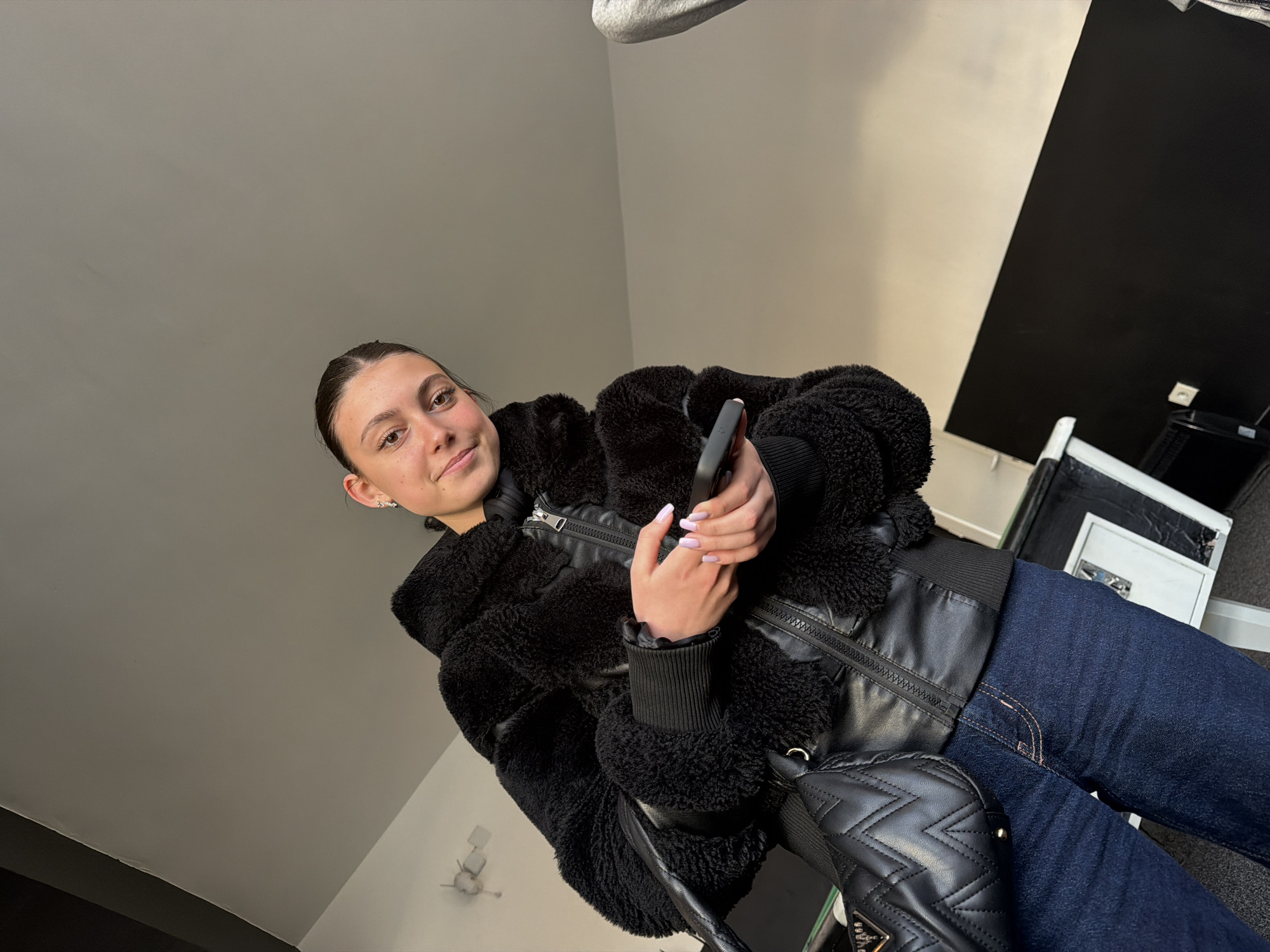 A woman is depicted in a low-angle shot, holding a mobile phone in her hands. She is wearing a black, fuzzy jacket with leather trim and dark jeans. A black quilted bag is slung over her shoulder. The background consists of a light grey ceiling and a wall that transitions from light grey to black. A white cabinet is visible in the background. The location information of Veliki Preslav, Bulgaria is not discernible from the visual elements of this image.