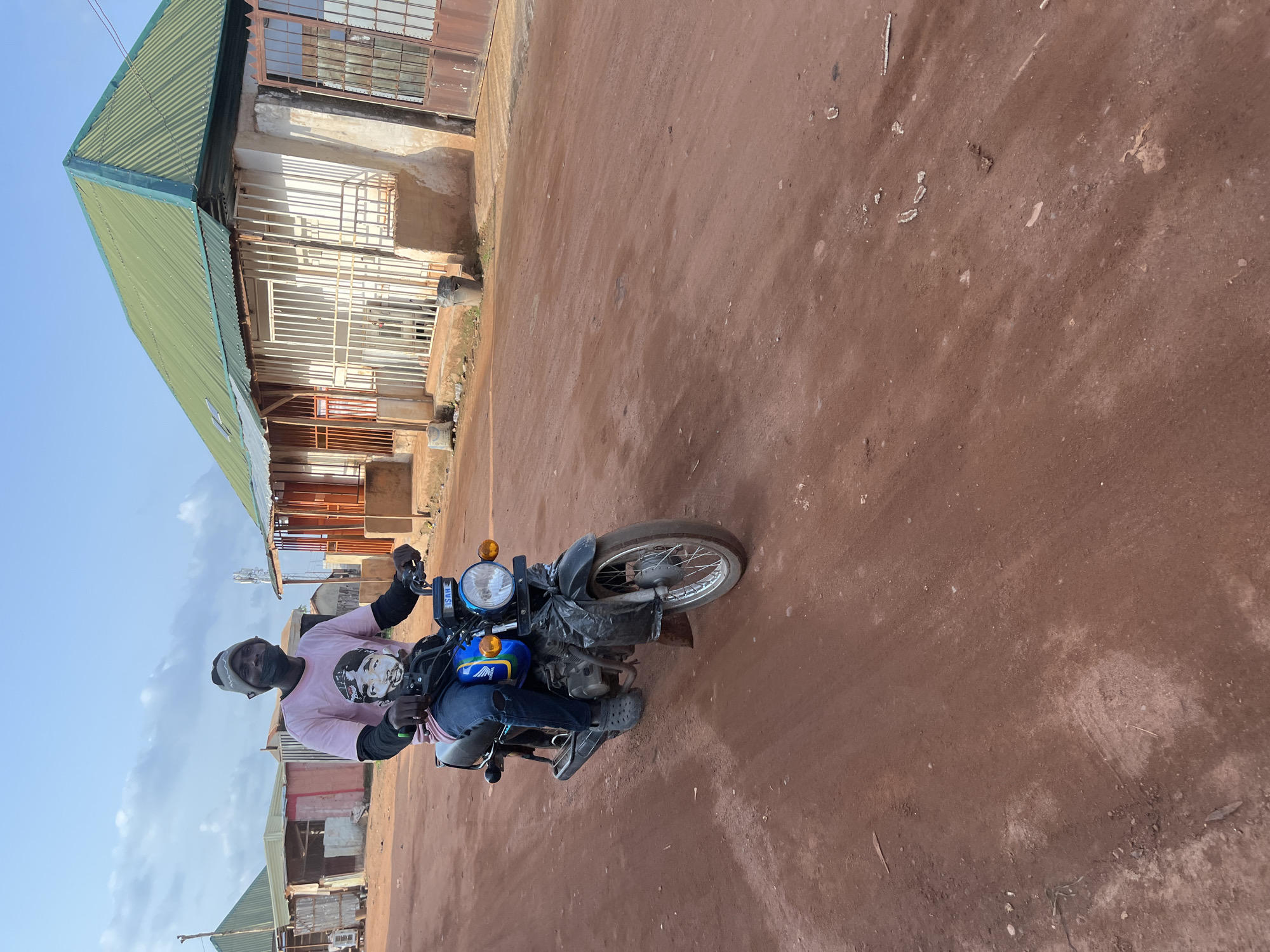 In Abuja, Nigeria, a Black man wearing a pink t-shirt with a portrait, blue ripped jeans, and a grey beanie rides a black motorcycle on a dirt road. The motorcycle has a blue fuel tank with a yellow and blue logo and a black plastic covering over the front fender. The man is looking towards the camera with a black face mask pulled down below his nose. Behind him, there are several buildings with corrugated green roofs and barred windows. The road surface is reddish-brown dirt with tire tracks visible. The sky is partly cloudy.