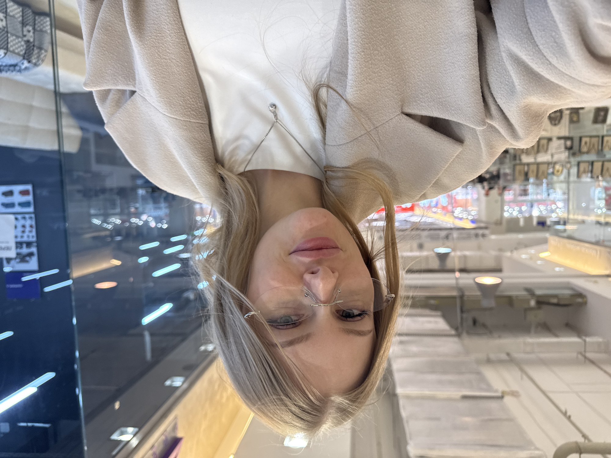A woman is photographed from an inverted perspective in what appears to be a retail environment. She has blonde hair, wears frameless glasses, and is attired in a white top and a beige coat. A delicate necklace is visible.
The background indicates a shopping center or department store interior with a display of items, possibly jewelry, and other retail fixtures. Reflections suggest glass surfaces and interior lighting characteristic of commercial spaces.
Without additional corroboration, definitively determining the exact location as Perm, Russia, based solely on the visual data, is not possible.