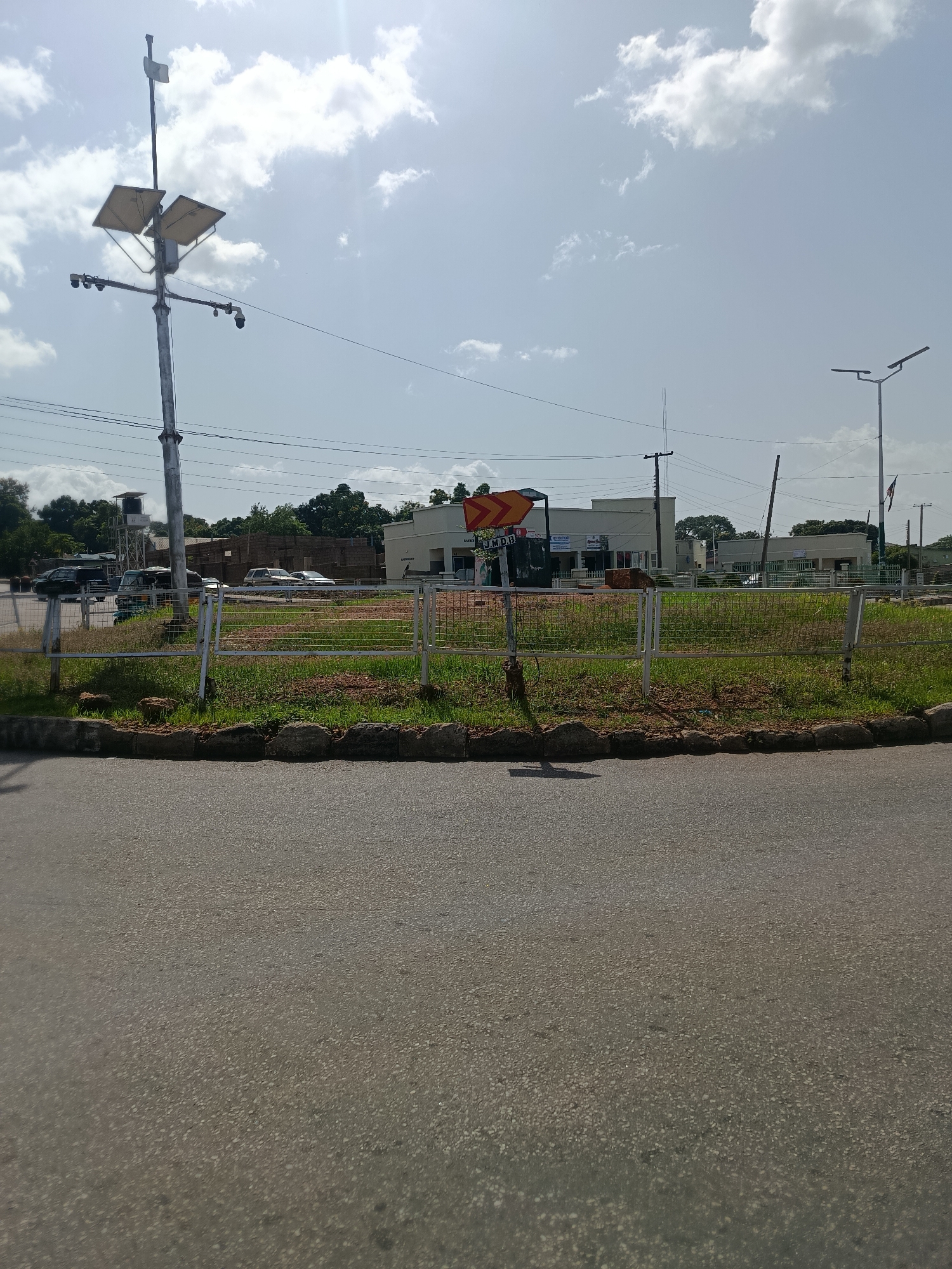 The image depicts a street scene in Jos, Nigeria, under a bright, cloudy sky. In the foreground, a paved road leads towards a grassy median strip with a white metal fence. Beyond the fence, a row of buildings lines the street, some appearing to be commercial establishments with signs that are difficult to read clearly. A tall metal pole with solar panels and what appear to be security cameras is prominent on the left side of the image. Several vehicles, including cars and a van, are parked or driving along the street in the background. Trees and vegetation frame the buildings. A traffic sign with orange and yellow arrows is visible near the metal pole. The overall impression is of a typical urban environment on a sunny day.
