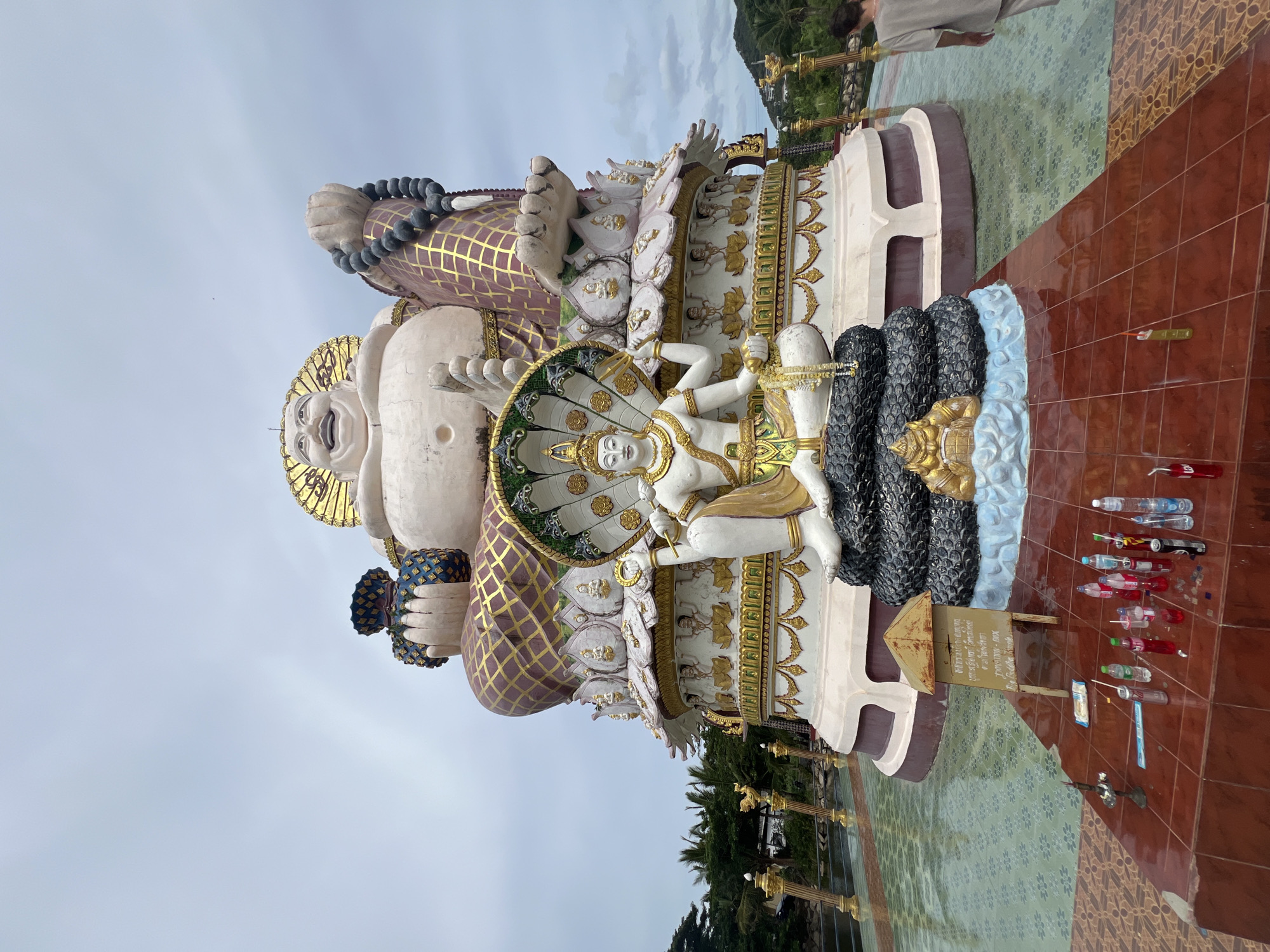 This media file depicts a large, colorful statue with multiple figures in an outdoor setting. The main subject is a reclining figure with a jovial expression, adorned with a golden headdress and a purple and gold patterned robe. Around its neck, it wears a string of large dark beads. Another figure, depicted as a deity with multiple arms and a serene expression, is seated and partially cradled by the reclining figure. This seated figure is surrounded by a multi-headed cobra hood, rendered in intricate detail with gold accents. The statues are positioned on a multi-tiered platform decorated with floral motifs and smaller figures.
In the background, lush green foliage and distant hills suggest a tropical location. The ground in front of the statue is tiled with a mosaic of green and brown patterns. Scattered on the tiled area are various objects, including several plastic bottles, some with colored liquid, and what appear to be red sparklers or incense sticks. A small sign with some writing is also visible near the base of the statue. The sky is overcast, indicating a cloudy day. The overall scene suggests a place of religious or cultural significance, possibly a temple or a tourist attraction. There is a partial view of a person in a light-colored garment standing near the top edge of the frame, and another person partially visible in the far upper right corner.
The writing on the sign is in a language that appears to be Thai. The context provided indicates the location is Baan Plai Laem, Thailand.