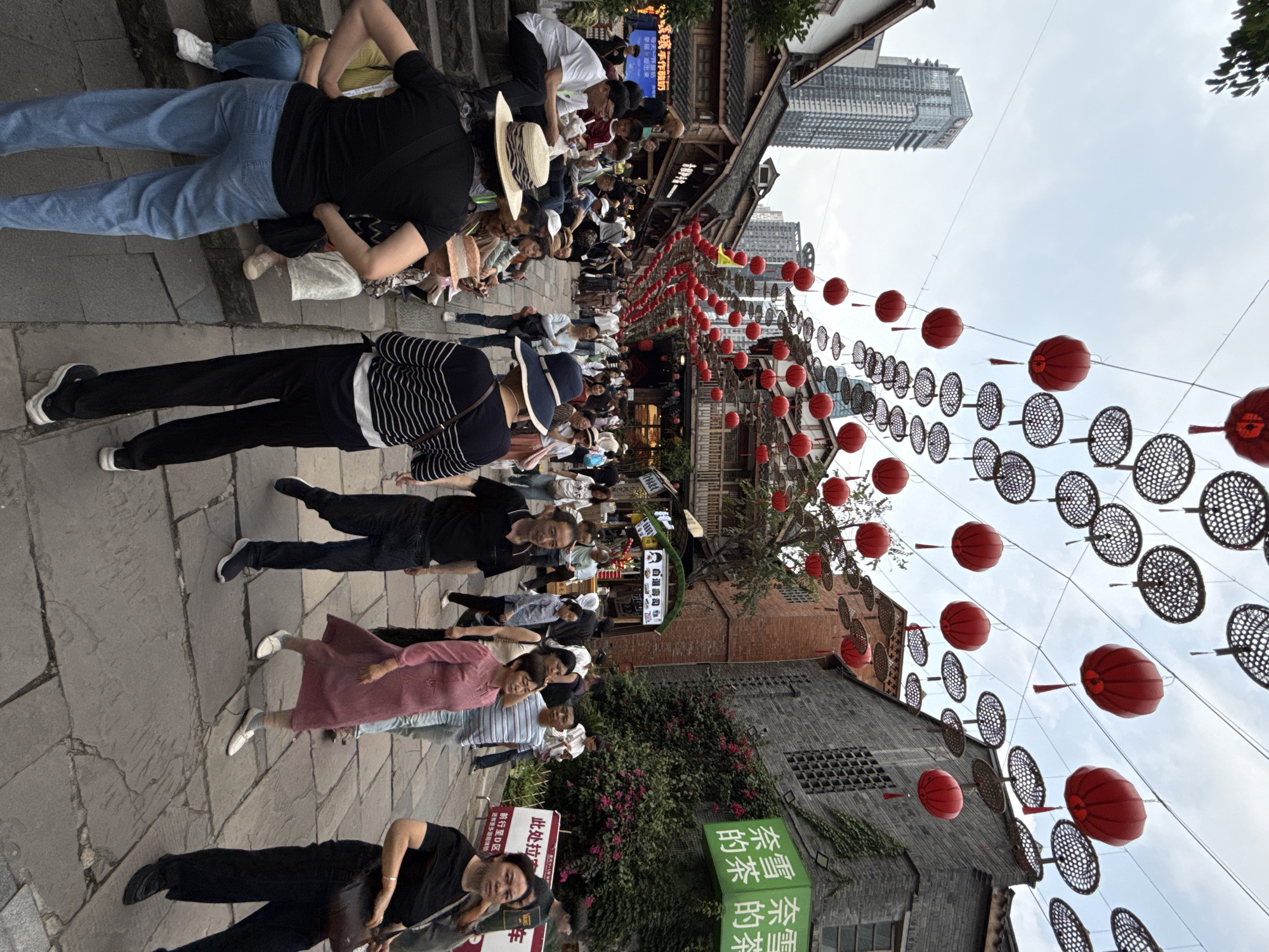 The image shows a street scene in what appears to be a commercial area of Yuzhong District, China. Red lanterns and decorative round screens are strung overhead. A crowd of people is present, with some seated or lying on the ground, and others walking or standing. Various signs with Chinese characters are visible, one appearing to advertise "奈雪的茶" (Nayuki's Tea). Buildings in the background display a mix of traditional and modern architectural styles.