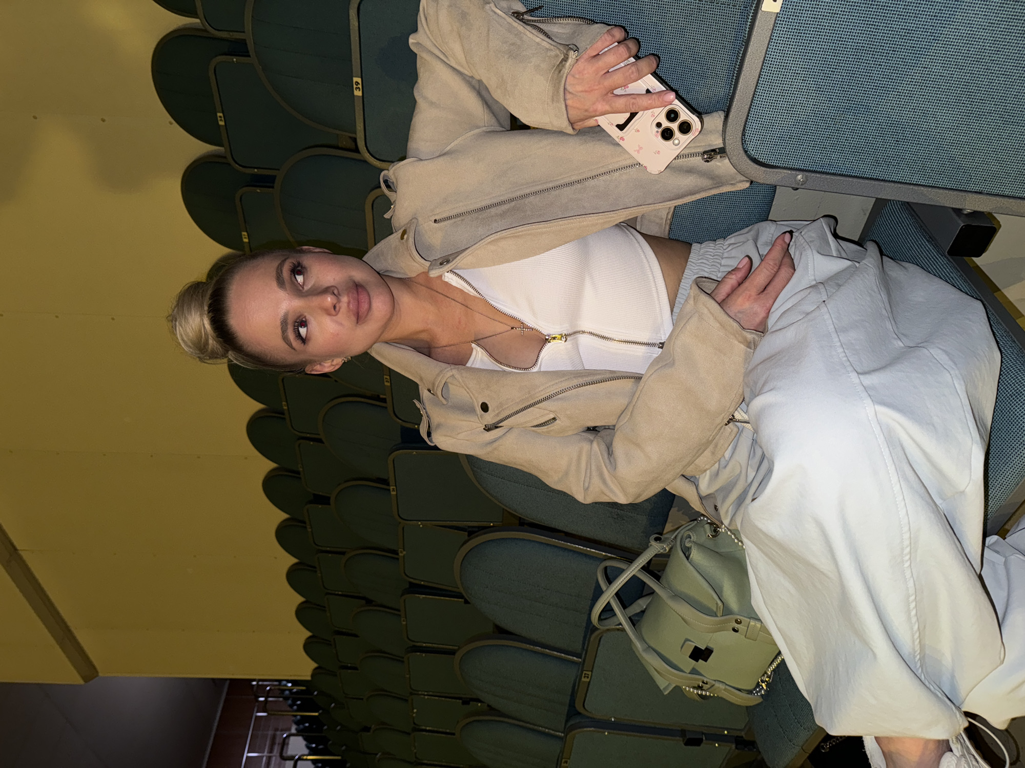 A young woman is seated in what appears to be an auditorium or theater. She has blonde hair tied in a bun and is looking directly at the camera with a neutral to slightly pouty expression. She is wearing a light-colored, possibly beige or light grey, zippered jacket over a white cropped tank top with a zipper. Her lower body is clad in light blue or light grey baggy pants.
Her left hand holds a smartphone with a light pink case that features a dark letter, possibly 'V' or 'A', and small pink floral or butterfly patterns. Her right hand is resting on her lap, partially hidden by her clothing. A light green handbag with chain details is visible on the seat or floor next to her feet.
The setting consists of rows of dark green upholstered seats, receding into the background. Some seats have visible numbers on their backs, including "39", "38", "37", "3", and "4". The wall to the left of the woman is a plain, light yellow or beige color. The overall lighting is dim, suggesting an indoor evening event or a low-light environment during the day.