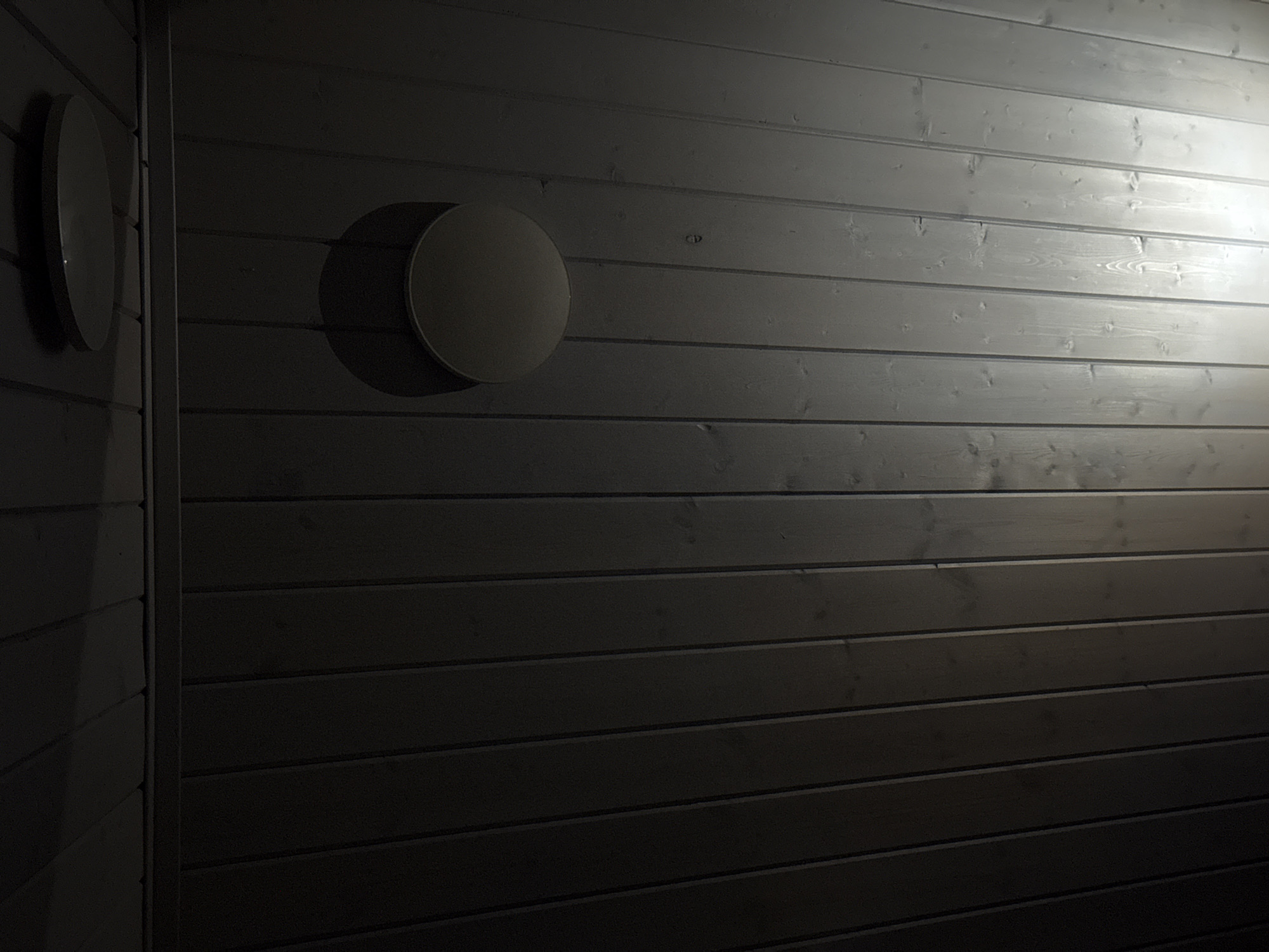 The image shows a dark wooden ceiling with two circular light fixtures. One fixture is on the upper left and is mostly in shadow, appearing as a dark circle with a faint highlight. The second fixture is on the right side of the image, also circular and illuminated, casting a bright light downwards. The ceiling planks are oriented vertically from the perspective of the image. The overall lighting is dim, with the exception of the area directly under the illuminated fixture. The location provided, Doma 1096 Km, Russia, is a postal code and likely refers to a remote or rural area, potentially a dacha or a standalone building. There are no people or visible actions depicted in the image.