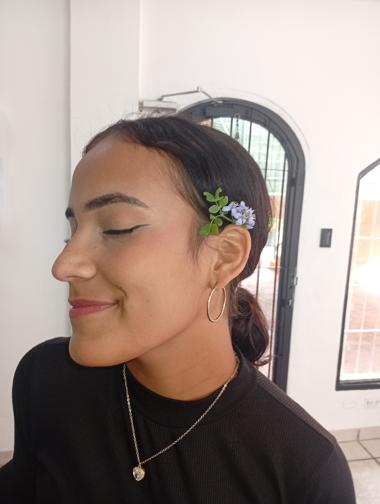 A young woman is seen in profile, indoors. She has dark hair pulled back, with small blue flowers and green leaves tucked behind her ear. She is wearing gold hoop earrings and a gold necklace with a heart-shaped pendant. She has a slight smile and her eyes are closed. She is wearing a black, ribbed long-sleeved shirt.
Behind her is a white wall with a black, arched door or window with a barred frame. Through the door or window, a glimpse of the outside can be seen, appearing to be a building and possibly a wall. The lighting is bright and appears to be from natural daylight.
There is no information in the image to indicate it was taken in Juan Griego, Venezuela.