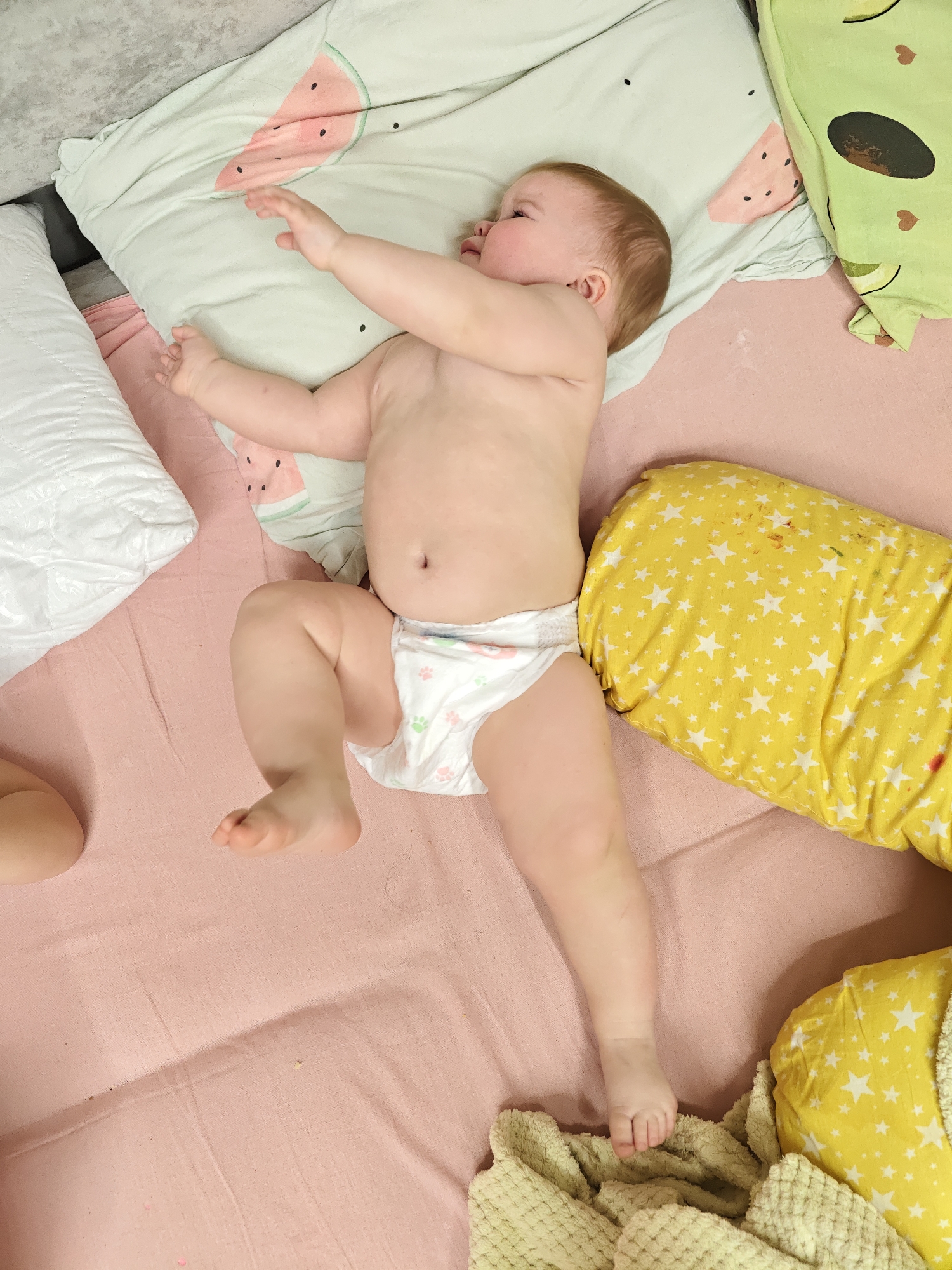 A baby wearing a diaper lies on a pink sheet, nestled amongst pillows and blankets with different patterns (watermelon, avocado, stars). The location is presumed to be Chaikovsky, Russia.