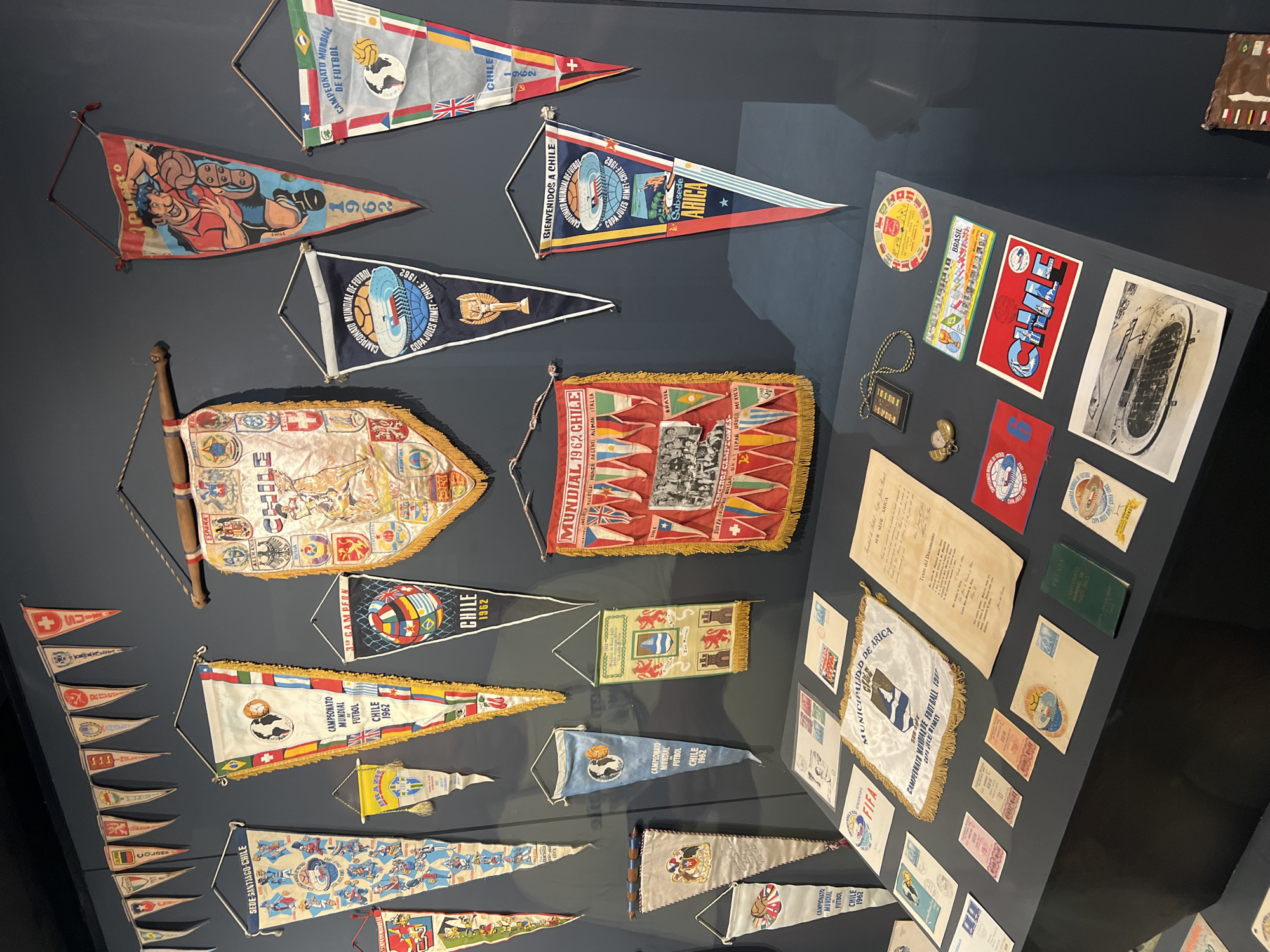 The image shows a display of memorabilia related to the 1962 FIFA World Cup, held in Chile. The display features numerous pennants, stickers, and documents related to the tournament. Several pennants are visible, bearing the names of participating nations like Spain, Germany, Argentina, Brazil, Yugoslavia, and Switzerland. Many items mention "Chile 1962," highlighting the event's location. One pennant references "Subsede Arica," indicating the city's role as a sub-venue. Other items include the "Copa Jules Rimet" (the World Cup trophy at the time), logos related to FIFA and the Municipality of Arica, and various printed documents. A number of the pennants advertise Arica as a location.