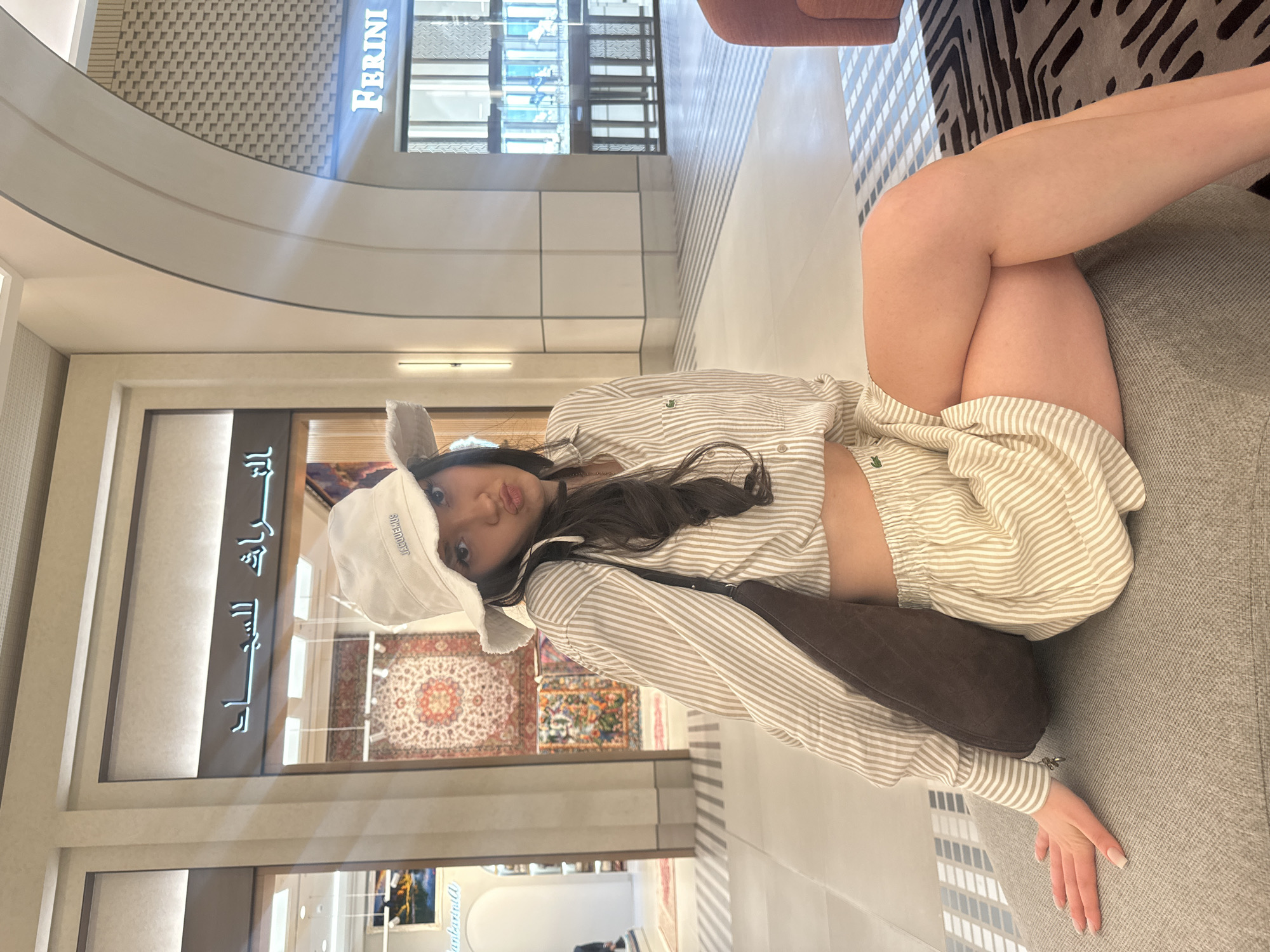 A young woman is depicted lounging on a light grey upholstered bench or couch, looking directly at the camera with her lips slightly pursed. The image is oriented sideways, but she appears to be relaxed, with one leg bent and crossed over the other.
She is dressed in a casual, light-colored ensemble, including a beige and white striped long-sleeved button-up shirt, which features a small green crocodile logo (Lacoste) on the left chest. She wears matching striped shorts. A dark brown shoulder bag is slung across her body and rests on the couch beside her. A white bucket hat with the brand name "JACQUEMUS" visibly stitched on the front covers part of her long, dark hair. Her fingernails are manicured with a light polish.
The setting appears to be a spacious, brightly lit indoor area, likely a modern shopping mall or commercial complex in Dubai, United Arab Emirates. The architecture features light-colored walls, curved ceiling elements, and patterned flooring. In the background, several storefronts are visible. A prominent blue illuminated sign overhead reads "FERRINI." Another store, partially obscured behind the woman, showcases colorful patterned rugs and features text in Arabic on its sign. Inside this store, another sign with partially visible English text, possibly "Van Der Burgh," can be seen. The overall environment suggests an upscale and contemporary commercial space, with bright, ambient lighting indicating it is daytime.