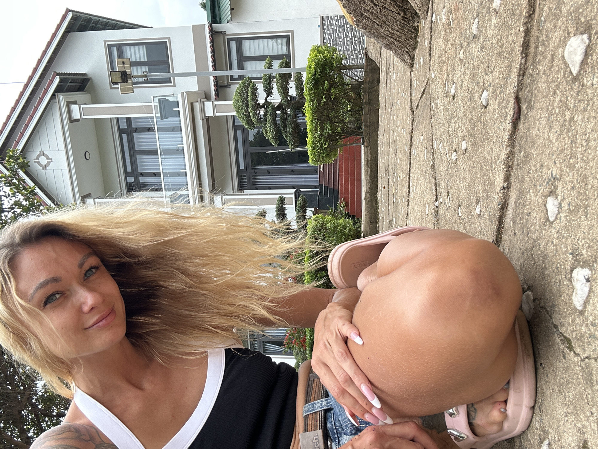A blonde woman with blue eyes and long, wavy blonde hair is crouching on a paved outdoor area in front of a modern multi-story house. She is wearing a black tank top with white trim, denim shorts, a belt, and pink slip-on sandals. Her left hand is resting on her leg, and her right leg is bent, with her knee pointing towards the camera. The house has a white facade with brown trim and a red tiled roof. There are windows, balconies, and a covered entrance with stairs leading up to a large glass door. In front of the house is a manicured garden with bushes and a decorative tree. The ground is paved with large concrete slabs, and there are some scattered white pebbles. The overall scene appears to be in a residential area in Thôn 2, Vietnam.