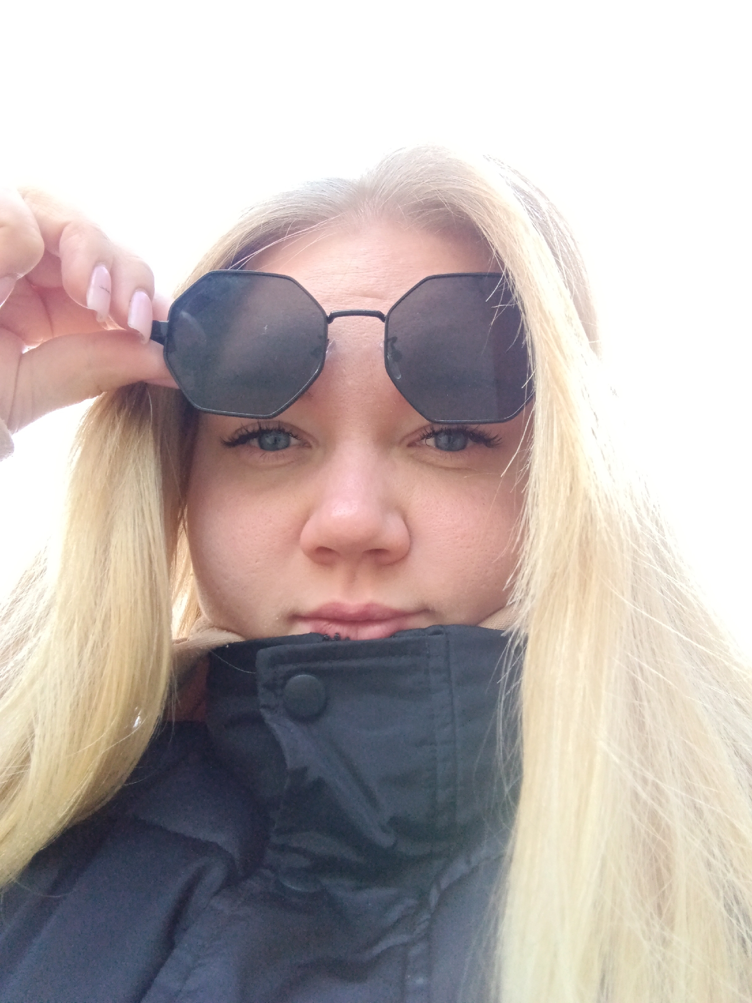 A woman is holding a pair of dark, octagonal sunglasses above her eyes. She has light blonde hair and is wearing a dark jacket with a buttoned collar. The background is bright, likely an overcast sky. There is no information in the image to definitively indicate the location is in Фоки, Russia.