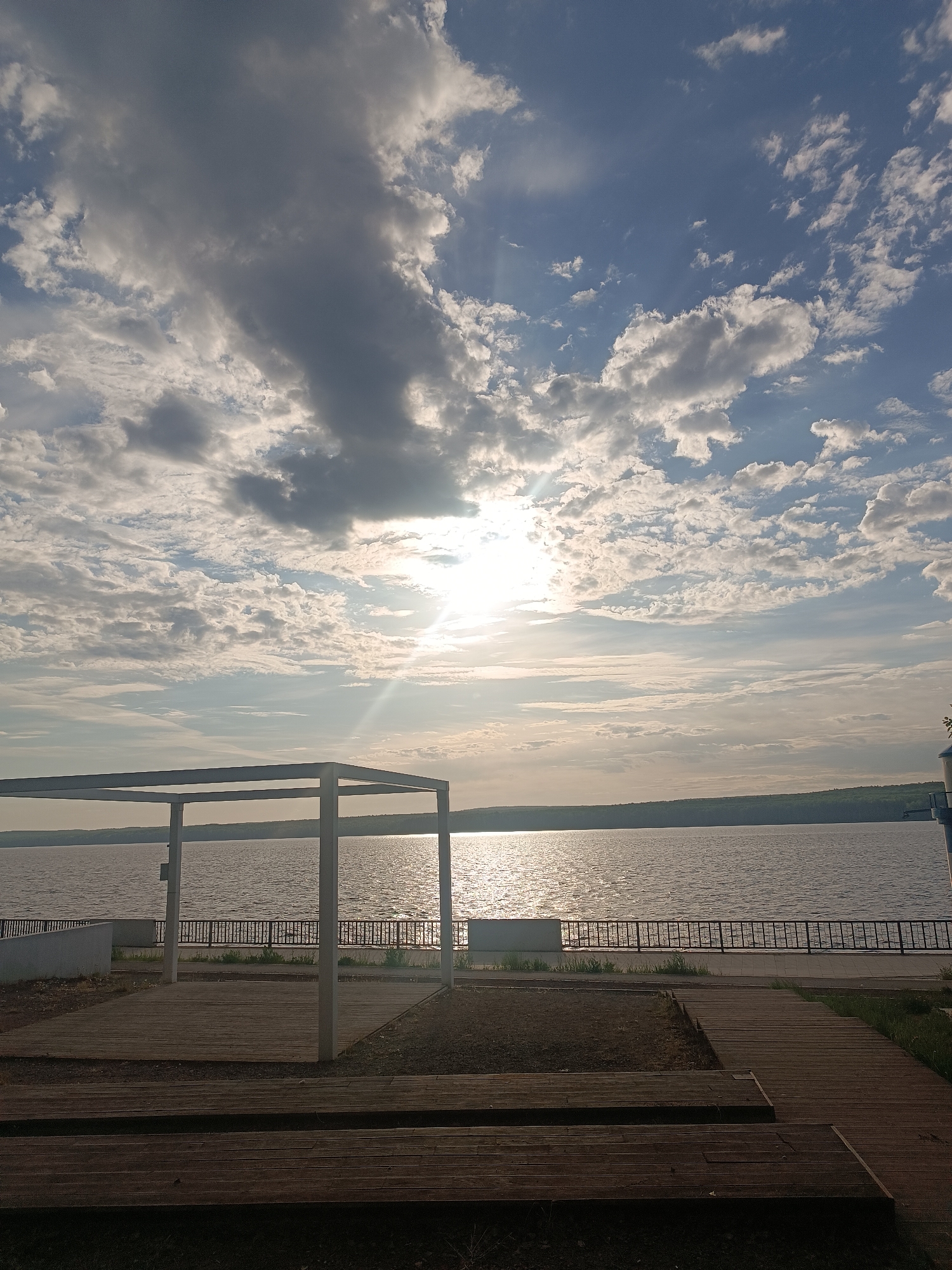 The image shows a view of the Kama River in Chaikovsky, Russia. In the foreground, there is a wooden boardwalk and a simple white metal structure. A metal railing lines the riverbank, separating the boardwalk from the water. In the background, the Kama River stretches out under a partly cloudy sky with the sun visible through the clouds. The opposite shore is visible in the distance. The time of day appears to be late afternoon or early evening based on the sun's position and the lighting. The overall scene conveys a sense of tranquility.