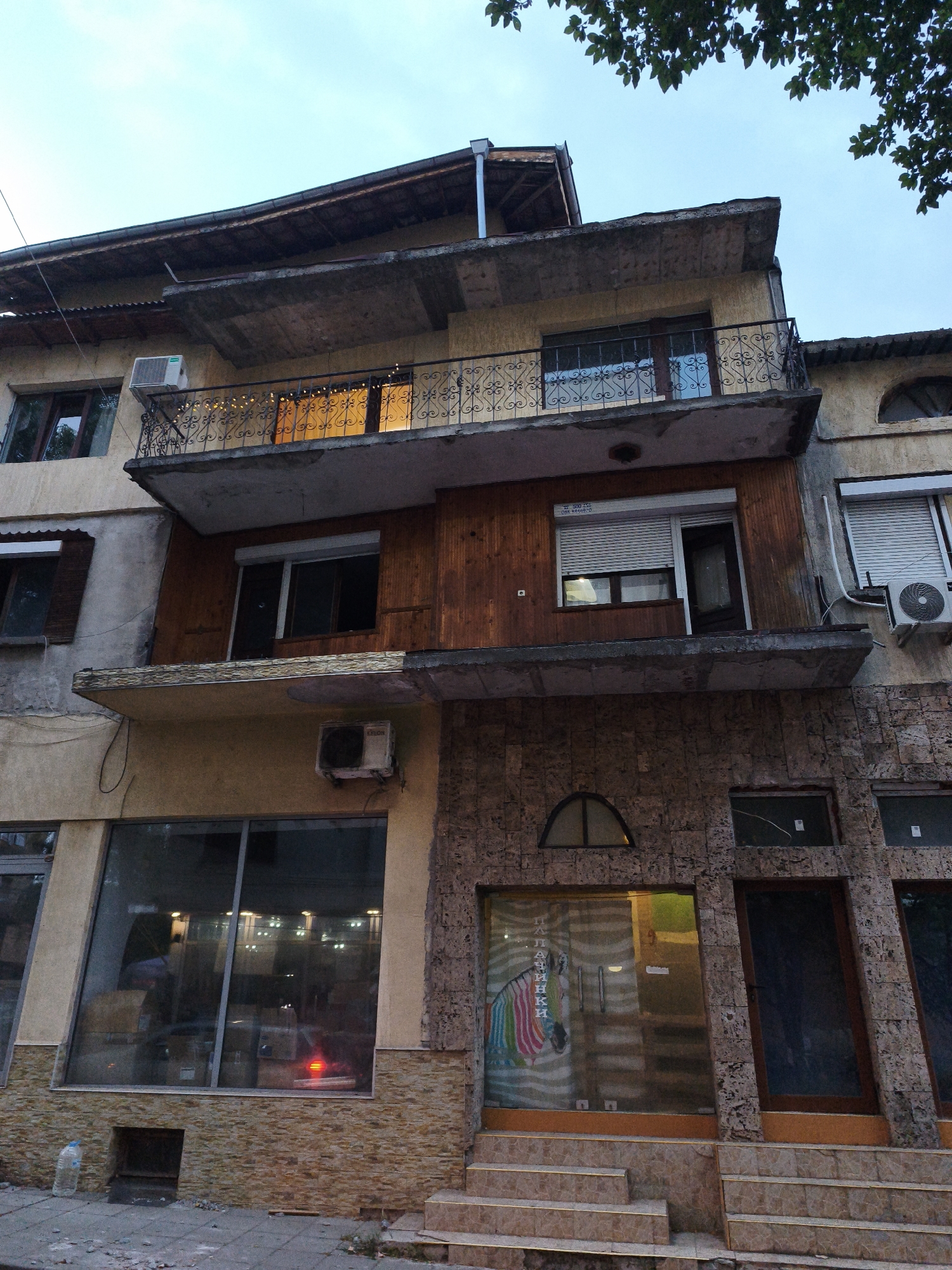 The image shows a multi-story building in Lovech, Bulgaria, with a mix of residential and commercial spaces. The facade is varied, featuring stucco, wood paneling, and stone elements.
On the upper floors, there are balconies with decorative railings, windows, and air conditioning units. One balcony has string lights. The roofline appears aged, with visible structural elements.
The ground level features large glass storefronts. One storefront displays a colorful image of a unicorn or horse with rainbow stripes and text in Cyrillic script, possibly indicating a business. Another storefront is a dark wooden door. Steps lead up to these entrances.
A water bottle is visible on the sidewalk in front of one of the ground-level openings. The overall impression is of a somewhat weathered but functional urban building, likely a mix of residential apartments above shops. The lighting suggests it is either early morning or late evening.