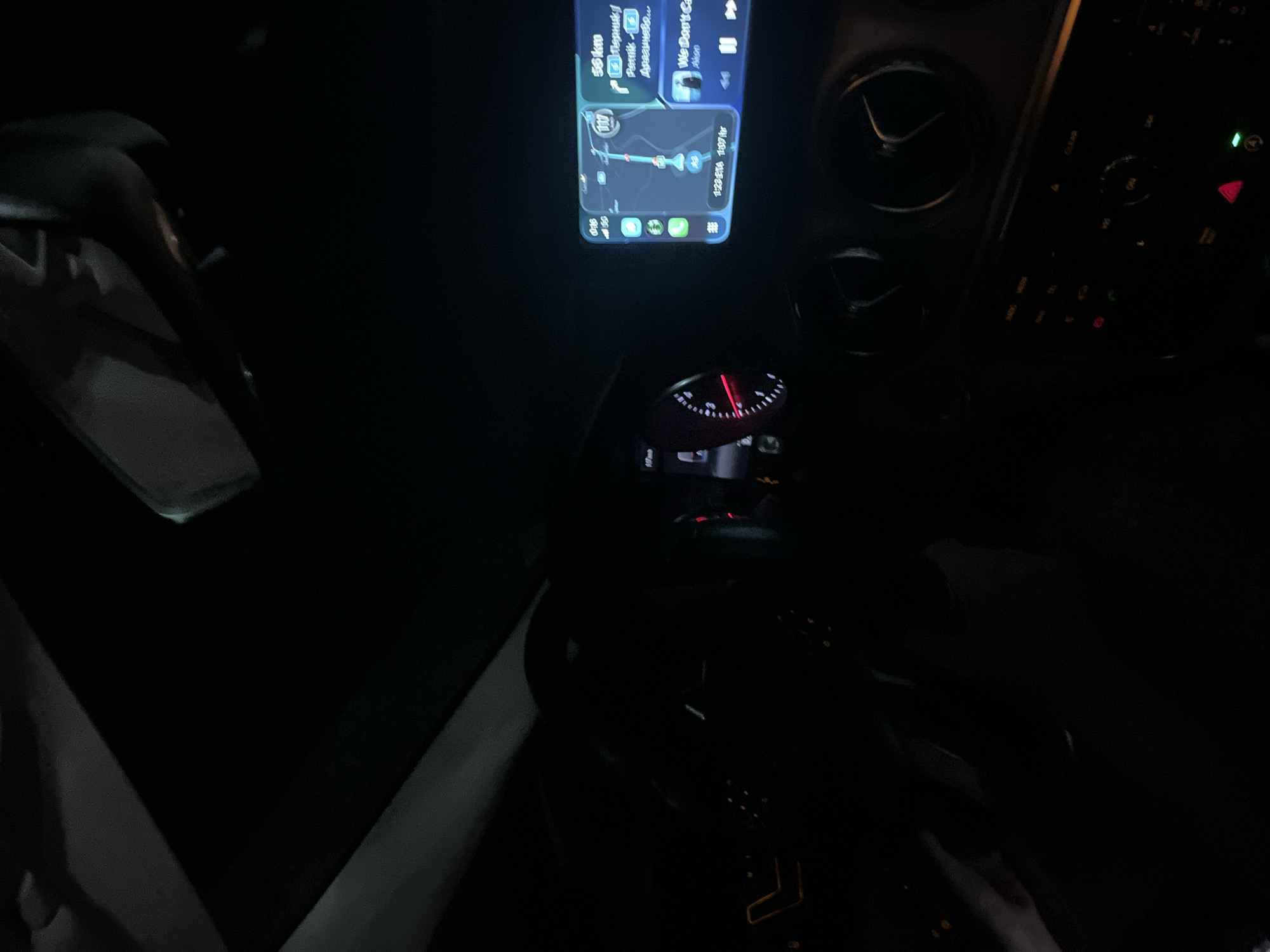 The image appears to be taken from inside a car at night. The dashboard is visible, with a phone displaying a navigation app mounted on it. The app shows a route indicating 56km to a location named "Перник" via routes numbered 116 and 117. The ETA on the navigation screen is 1:23 for an arrival time of 1:07. The speedometer needle is barely visible, but it shows a low speed. To the right of the phone are air vents and buttons of the car's infotainment system. The environment outside is dark, and the car is on a road or street. The location is in relation to the city of Pernik and a destination of “Anaaunolo”, both likely near Mursalevo, Bulgaria, as that is the implied current location. There are no visible people or interactions taking place.