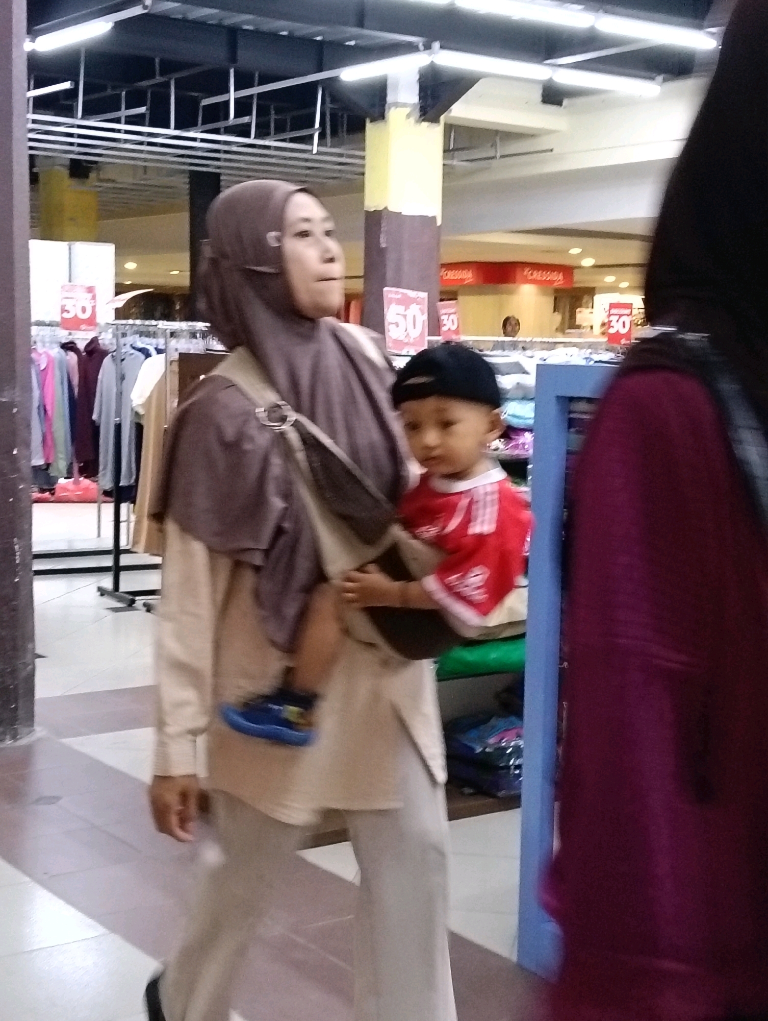 A medium shot captures a woman wearing a brown hijab and a light brown long-sleeved tunic, with a baby carrier slung over her shoulder. The baby, wearing a red shirt with white lettering and a black baseball cap, is visible in the carrier. The woman's legs are crossed, and she is wearing light gray pants and dark shoes. To the right, a portion of a person wearing a dark purple garment is visible. The background features clothing racks with various garments, signs displaying "50" and "30", and a light-colored tiled floor. The scene appears to be inside a store in Jaten, Indonesia, indicated by the retail environment and signage.