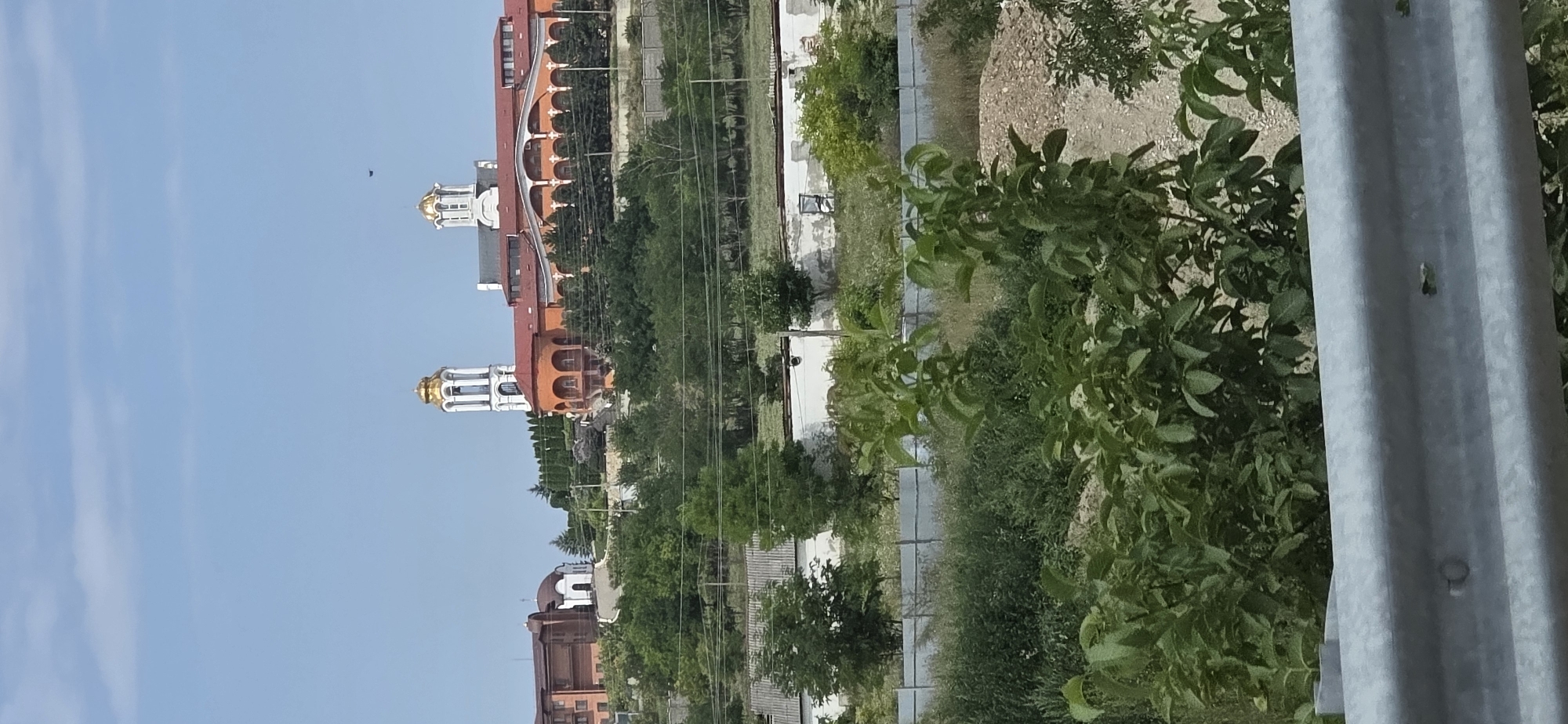 The image depicts a religious complex on a hillside under a clear blue sky. The main building features an orange facade with arched windows and a red roof. To its left, there is a white building with a small dome and a steeple. Further to the left, a taller brown building is visible. The complex is surrounded by lush green trees and vegetation. In the foreground, there is a metal barrier, possibly part of a road or viewing platform, with some green foliage in front of it. The overall scene suggests a serene and architectural setting. The location is identified as being in the city of Yasnaya Polyana, Russia.