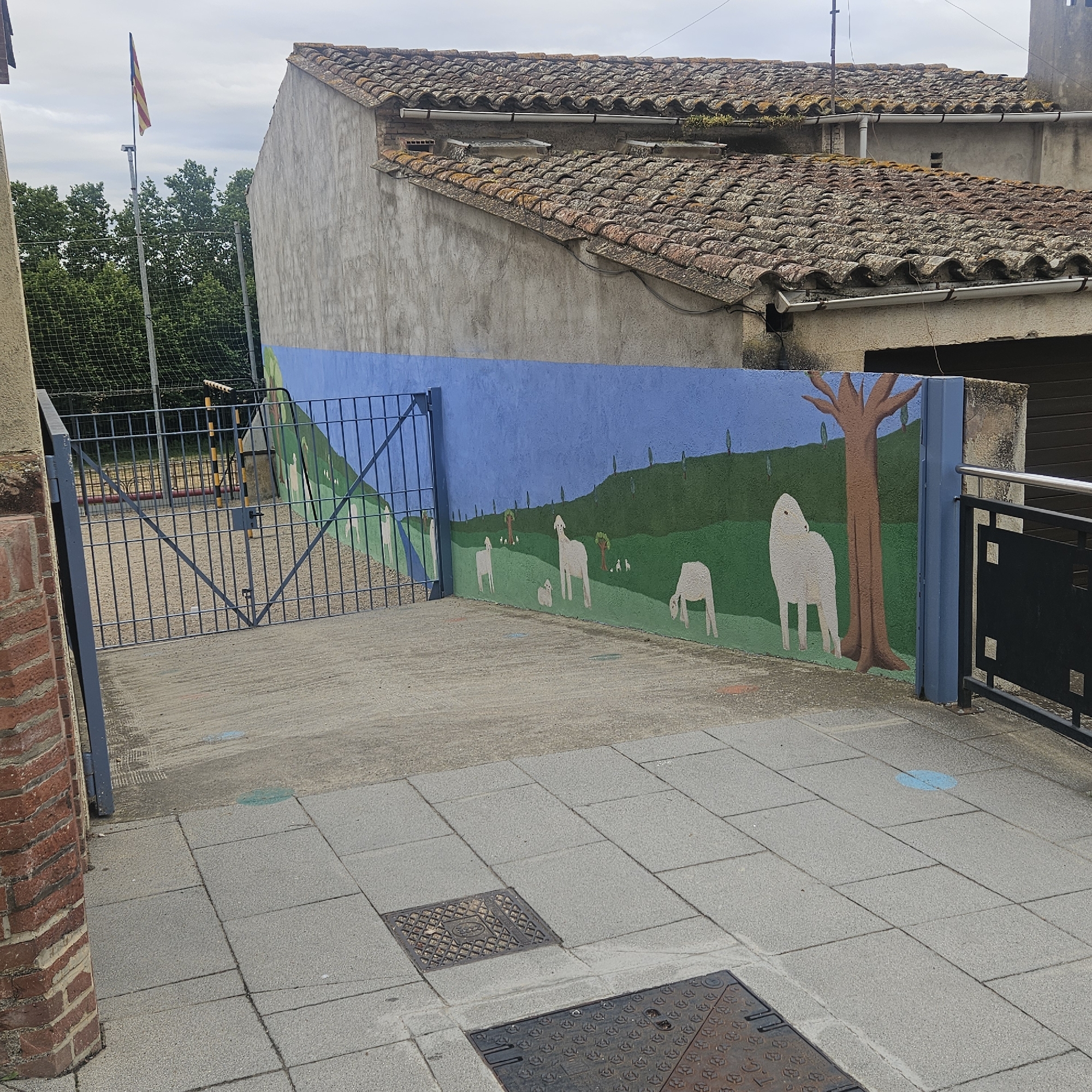 The image depicts an outdoor scene in Aiguaviva, Spain, featuring a mural on a blue-painted wall. The mural depicts a pastoral landscape with rolling green hills and scattered trees under a blue sky. Several white, stylized sheep are painted on the hills, some grazing and others standing. In the foreground, a metal gate with diamond-shaped openings provides access to a paved area. The gate is attached to a brick pillar on the left. Behind the gate, a chain-link fence encloses a grassy or dirt area, with a sports field visible beyond. A flag pole with a striped flag, possibly a Catalan flag, stands to the left of the fence. The ground in front of the mural and gate is paved with concrete tiles. In the background, a building with a red-tiled roof and stucco walls is visible. There are no people present in the image. The overall impression is one of a decorative element within a public or semi-public space, possibly a schoolyard or park.
