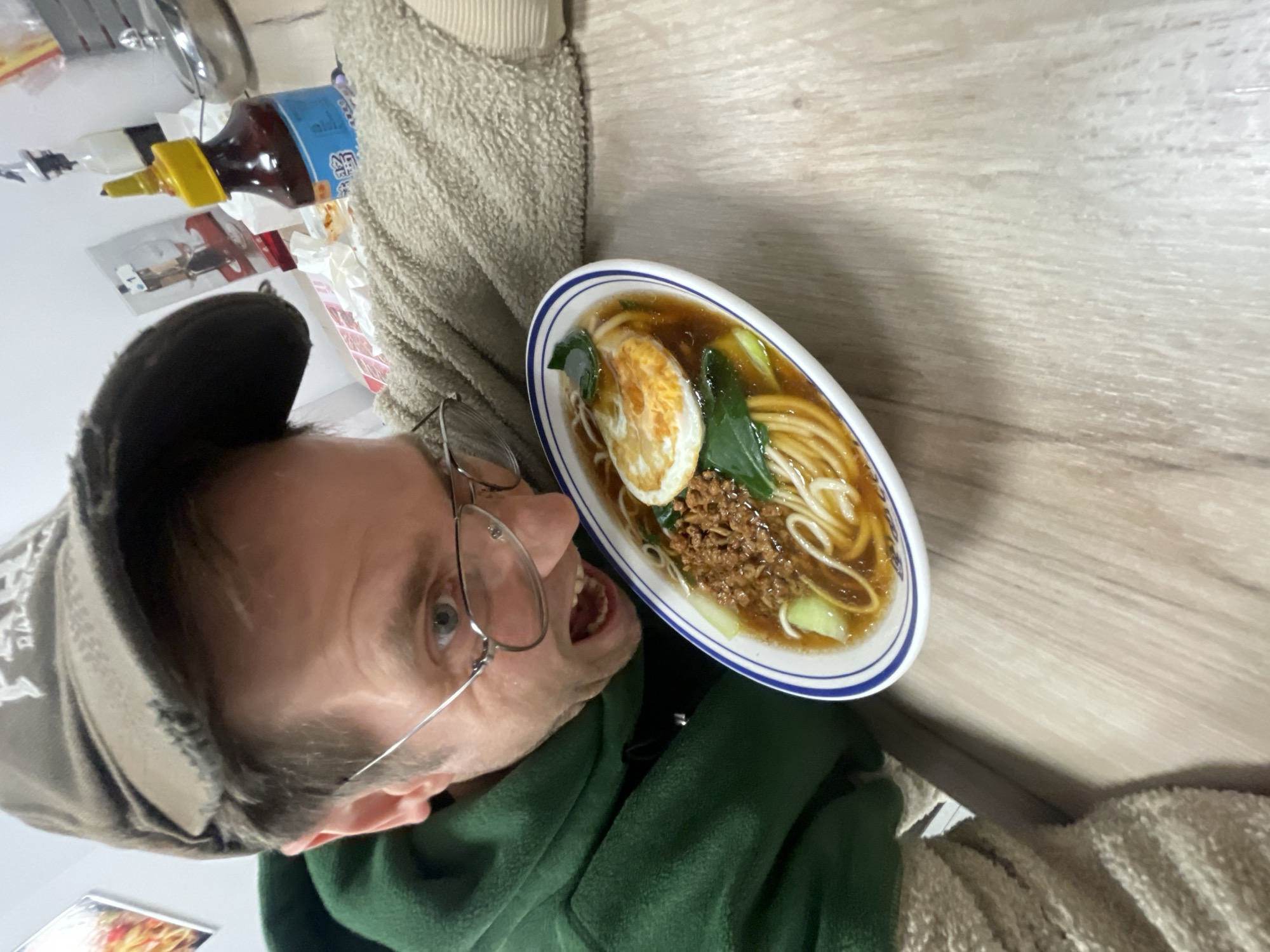 A man in a green hoodie and cap, wearing glasses, is holding a bowl of ramen. The bowl is white with a blue rim and contains noodles in broth with a fried egg, ground meat, and green vegetables. The background is a light brown wooden surface. A bottle of hot sauce and other items are visible in the upper left corner. The photo appears to be taken indoors and is rotated. There is no information to suggest this photo was taken in Belgrade, Serbia.