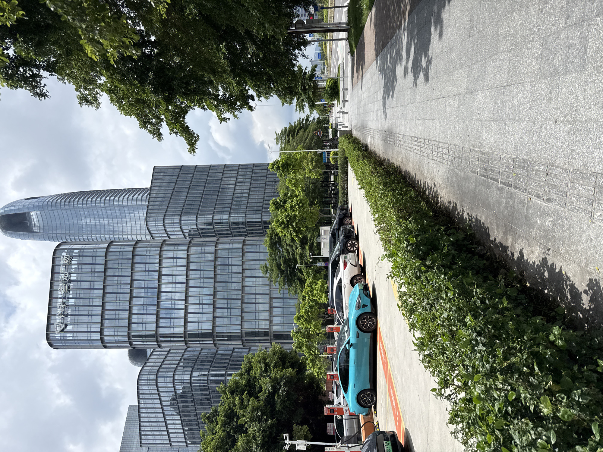 A wide, paved sidewalk runs along a row of dense green bushes and a parking lot in front of modern glass-faced office buildings in Haizhu District, China. The sidewalk is paved with large gray tiles and features a tactile paving strip for the visually impaired. Several electric cars are parked in the lot, with charging stations visible. In the background, several multi-story office buildings with reflective glass facades dominate the skyline. Tall green trees line the street and the parking lot. The sky is cloudy.