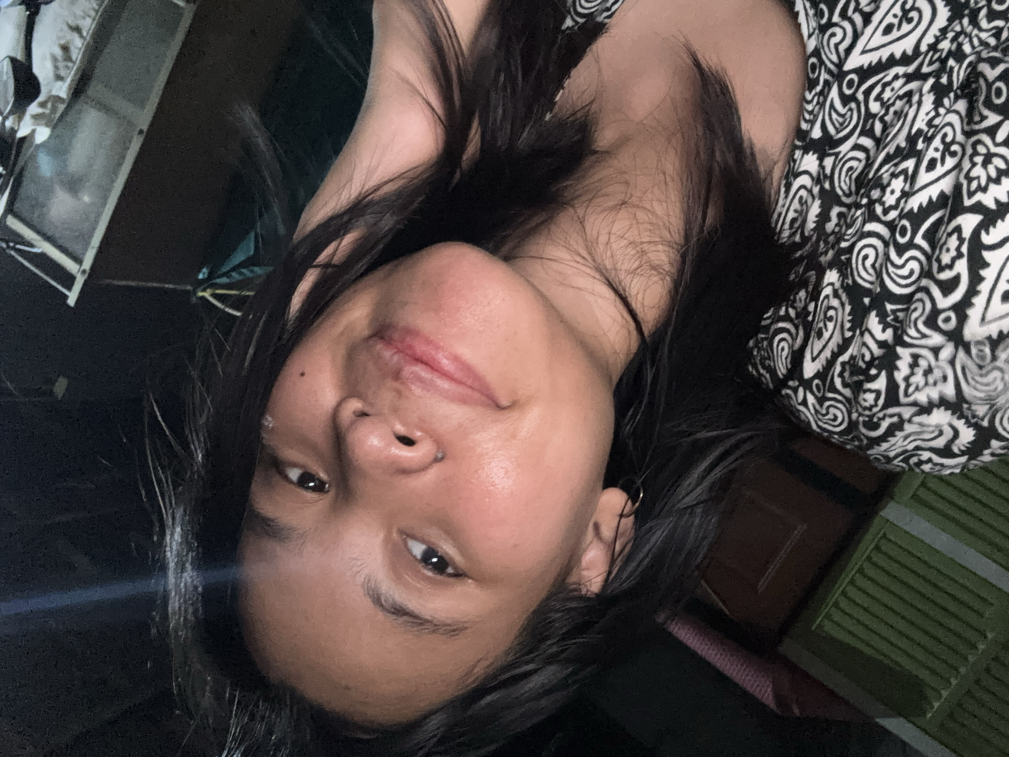 The image is a close-up selfie of a young woman with dark hair and light brown skin. She is looking towards the camera with a slight smile and her right hand is touching her hair. She is wearing a black and white patterned garment. The background is dark and appears to be indoors, possibly a bedroom, with some furniture visible. There is a bright light source illuminating the woman from the front, creating highlights on her face and hair. The overall impression is of a casual, personal photograph taken at night. There is no specific indication of location in East Jakarta, Indonesia, within the image itself.