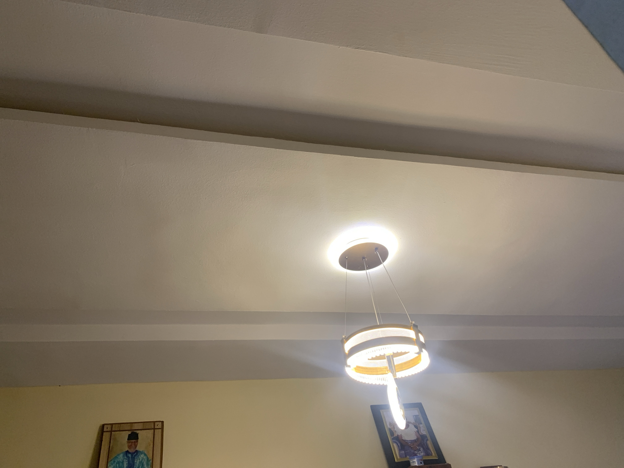 The image depicts an indoor scene with a modern chandelier hanging from a textured ceiling. The chandelier features multiple circular tiers illuminated by warm LED lights. Two framed pictures are visible on the wall below the ceiling. One is a portrait of a person wearing traditional attire, and the other is partially obscured. The environment appears to be a residential or commercial interior space. The exact location in Rido, Nigeria is not identifiable from the visual information provided.