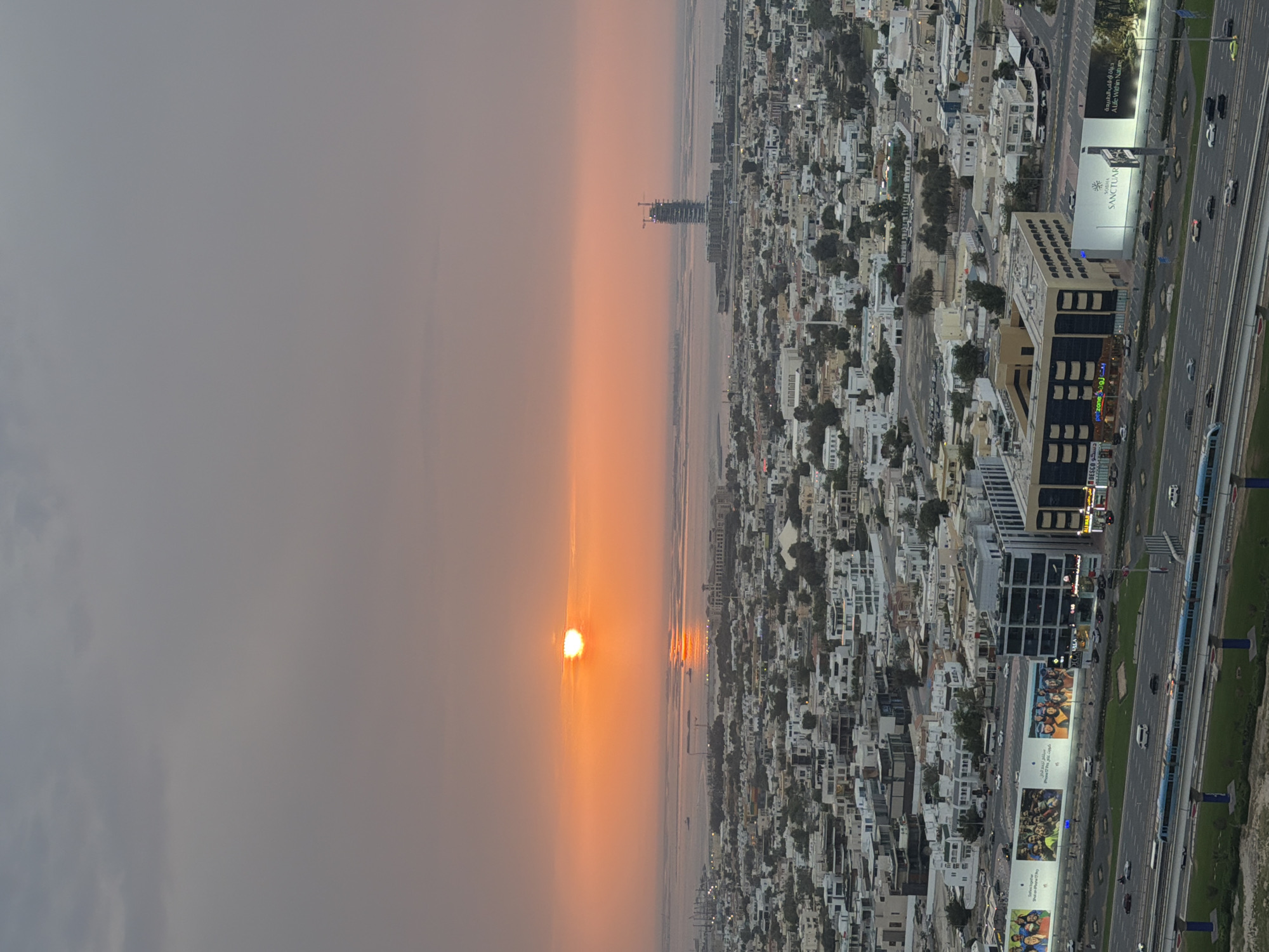 This high-angle, panoramic photograph captures a vibrant sunset over a bustling city, likely Dubai, UAE, given the architectural style and visible text. The sun, a brilliant orange disc partially obscured by atmospheric haze, dominates the left side of the frame, casting a warm, diffused glow across the sky. Its light reflects off a calm body of water, likely the Arabian Gulf, which stretches across the horizon below the sun. A distant, skeletal building under construction is visible on the far horizon, silhouetted against the bright sky.
To the right, a sprawling urban landscape unfolds with countless buildings, mostly light-colored and of varying heights, interspersed with green patches. Several multi-lane roads snake through the city, teeming with vehicles. A prominent elevated train or tram track runs parallel to one of the major highways, where a metro train is visible. Large commercial billboards line the highway, one clearly displaying "SANCTUARY" in English alongside Arabic script, and others showing advertisements featuring groups of people. A building nearby also has "ZONE" visible in its signage, accompanied by Arabic text. The scene conveys a sense of rapid urban development set against a dramatic natural phenomenon.