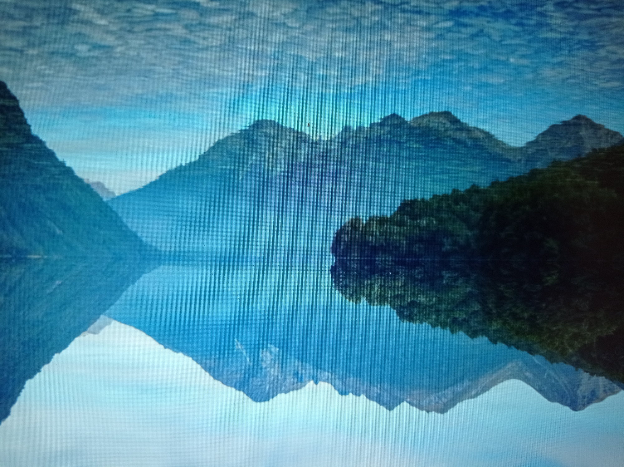 The image shows a serene natural landscape with a calm lake reflecting the surrounding mountains and a lush green forest. The mountains in the background are majestic and blue-toned, with some rocky patches visible on their slopes. The forest, located on the left side of the image, is dense and dark green, appearing to descend towards the lake. The water is crystal clear, revealing the rocky bottom of the lake in the foreground. The reflection of the mountains and forest is perfectly mirrored on the water's surface, creating a symmetrical and peaceful scene. The sky is a pale blue with some wispy clouds. This image does not depict any part of the city of Mukachevo, Ukraine.
