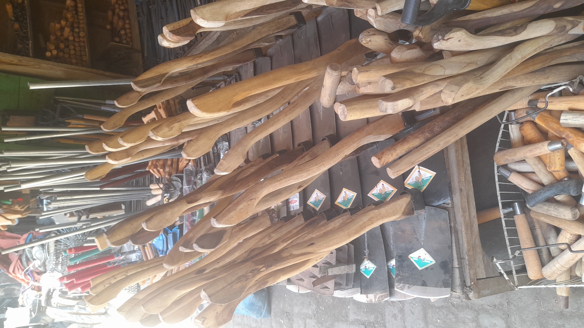 The image displays a street stall in Pekalongan, Indonesia, selling an assortment of agricultural tools, primarily hoes and their wooden handles. Numerous hoes with black metal blades are stacked on a wooden pallet, each fitted with a curved, light-brown wooden handle. These handles, varying slightly in shape and size, are the most prominent objects, filling the foreground and extending into the mid-ground. Behind the hoes, an array of long, metallic poles stands upright, likely more tool components or parts of the stall structure. To the right, shelves are visible, laden with more wooden dowels or handles. In the lower part of the image, a metal basket overflows with various wooden handles of different lengths and thicknesses, some with metal ferrules. The overall scene suggests an outdoor market setting, with the ground appearing to be concrete or dirt. The lighting indicates it is daytime.