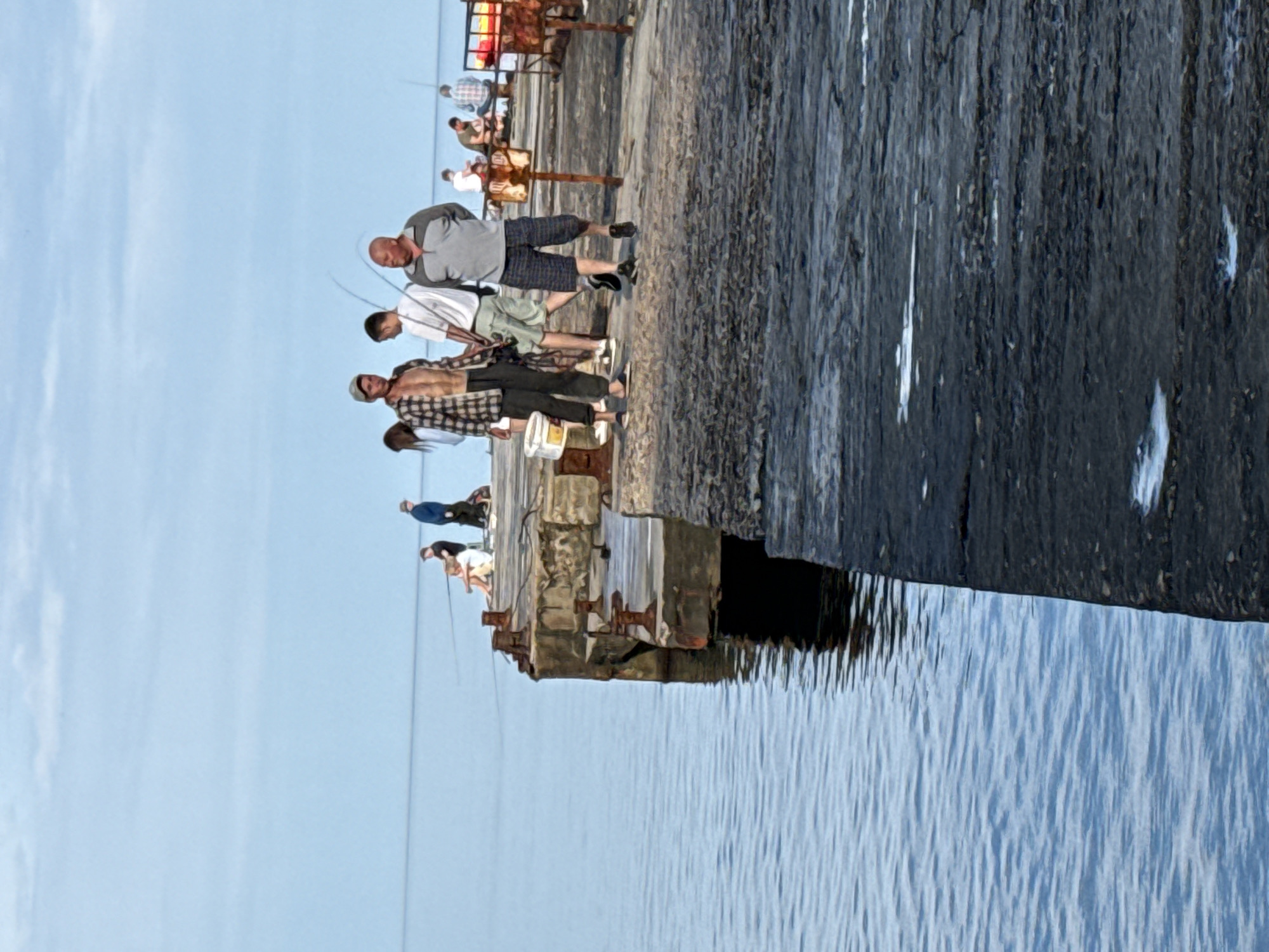 On a concrete pier extending into calm, blue water, several people are fishing. In the foreground, three men are walking and standing. The man on the left wears an unbuttoned plaid shirt over his bare chest and dark pants. Next to him, a younger man in a white t-shirt and light shorts walks. A bald man in a grey polo shirt and checkered shorts walks to the right of the younger man, looking forward. Further down the pier, several other individuals are seated and standing, some with fishing rods. The sky is a pale blue with scattered clouds. The pier has weathered concrete structures and metal fixtures.