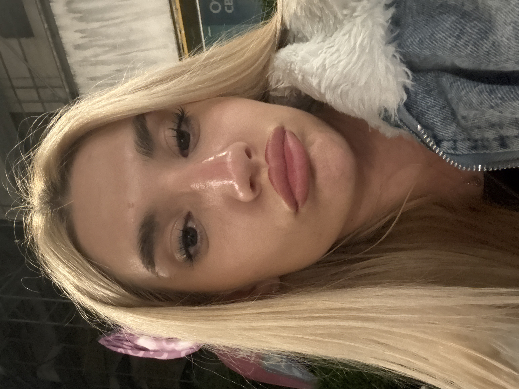 The image displays a close-up, rotated portrait of a young woman, featuring her head and upper torso. Her long, blonde hair cascades around her face. She is wearing a denim jacket with a prominent white, fluffy faux-fur lining around the collar. Her face is well-lit, highlighting her full, glossy pink lips, defined eyebrows, and long dark eyelashes with mascara. Her skin appears smooth and has a luminous, slightly shiny finish, particularly on her nose and chin.
The background is dark and out of focus, suggesting the photo was taken indoors or at night. Partial, blurred text, including fragments like "O" and "CE," is faintly visible on a background element, possibly a sign or poster. A glimpse of a purple and pink patterned fabric can be seen near the bottom left edge of the frame. The subject's expression is neutral as she looks directly at the camera. The overall impression is a stylish portrait taken in low-light conditions, possibly in Sofia, Bulgaria, based on the provided context.