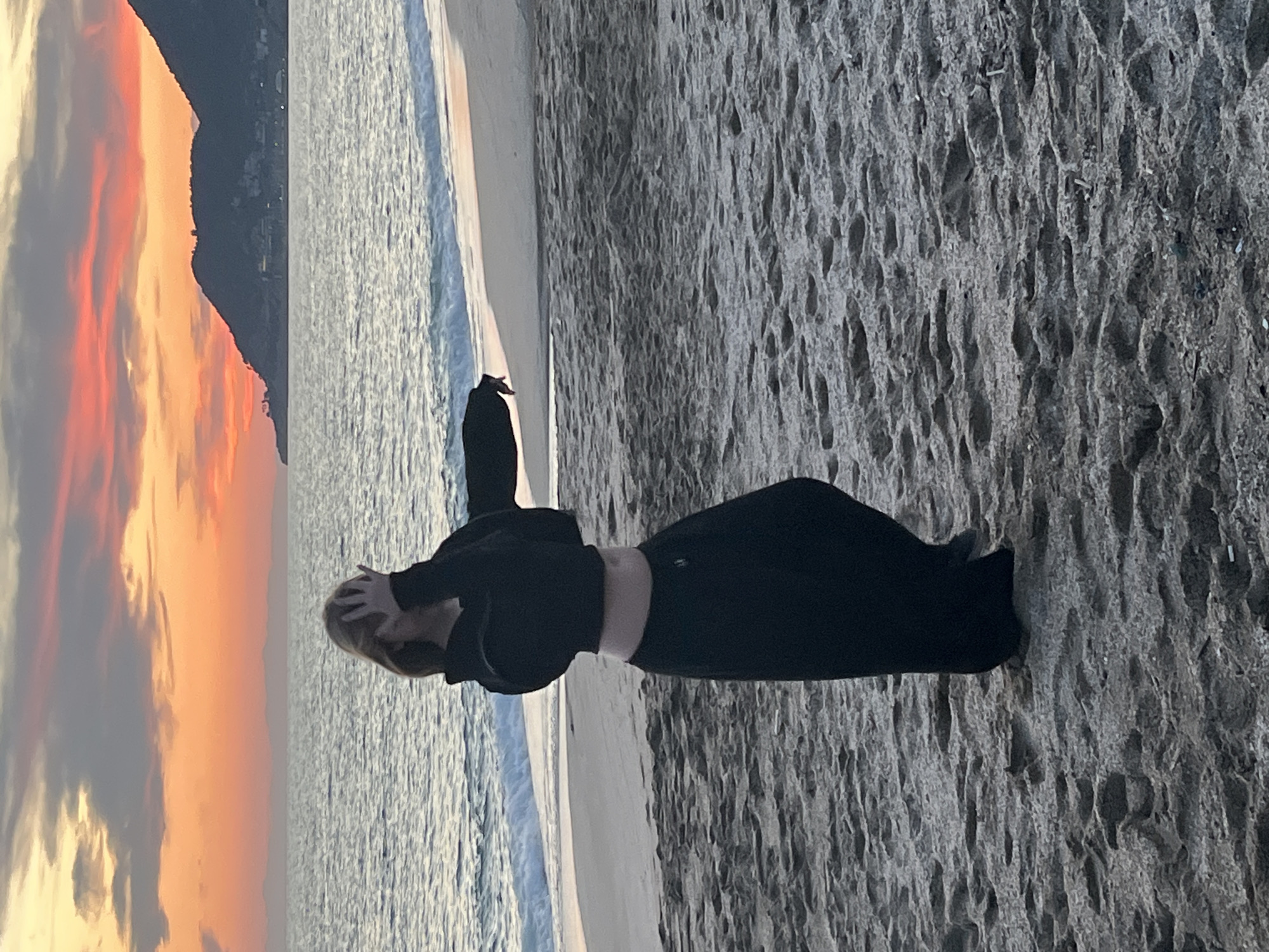 An outdoor medium shot captures a serene beach scene at either sunrise or sunset in Alanya, Turkey. The main subject is a person, likely female, with blonde hair, standing on the wet, textured sand with their back partially to the viewer. They are dressed in an all-black ensemble, consisting of a long-sleeved top or hoodie and full-length pants, with a sliver of their midriff visible. Their hands are placed on or near their head, suggesting a contemplative or relaxed posture as they face the ocean.
Beyond the person, the calm ocean stretches out, with gentle waves noticeable near the shore. The sky above the water is dramatic and vibrant, filled with clouds painted in rich hues of orange, red, and yellow, indicative of the early morning or late evening light. In the distance, a dark silhouette of a mountain range or large hill rises against the colorful sky, emphasizing the coastal setting. The sand in the foreground is patterned with ripples and textures, likely from receding waves. No visible text is present in the image.