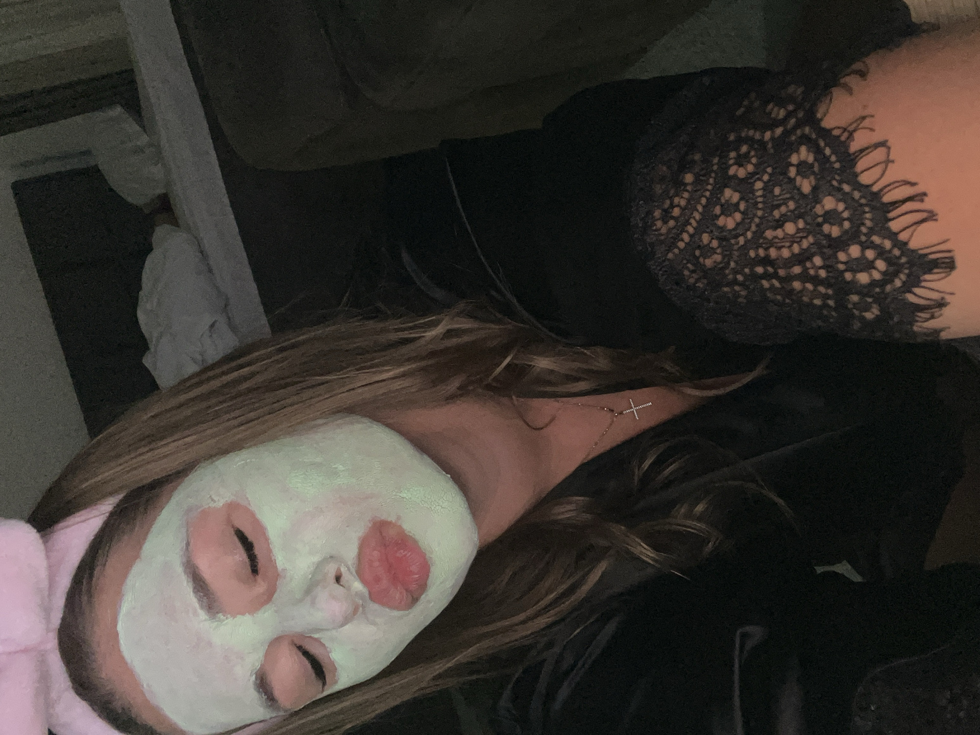 A woman with light brown hair is lying down, wearing a pink headband and a light green face mask with pink accents around her eyes and lips. She is wearing a black top with lace detailing on the shoulder. She has a silver necklace with a cross pendant. The background includes a light-colored wall or structure and dark fabric. The location is not discernible from the image.