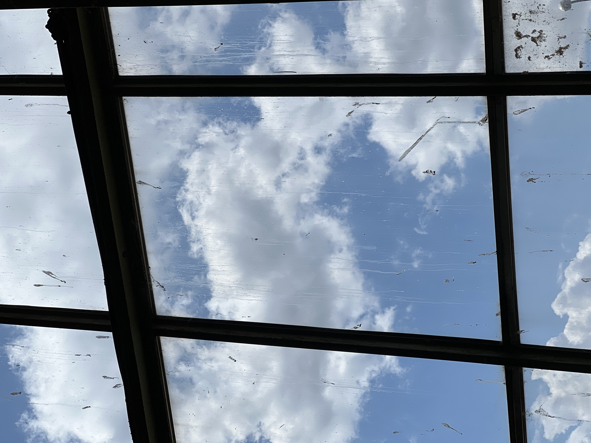 The image shows a view of the sky through a dirty, multi-paned window with a dark frame. The sky is blue with fluffy white clouds. Streaks and smudges, possibly from rain or dirt, are visible on the glass. Some of the streaks have dried into irregular patterns. The window is divided into several rectangular sections by thick, dark mullions. In the upper right section, a faint reflection or image of what appears to be a construction crane is visible against the clouds. The overall impression is of an aged, possibly industrial or neglected window looking out onto a bright day. The location is the city of Lanžhot, Czechia.