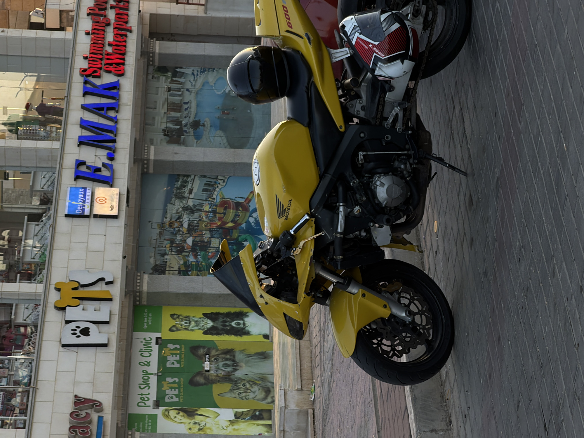 A yellow Honda sportbike is parked on a brick-paved sidewalk in front of a building in Sharm El Sheikh, Egypt. The building has large windows with a mural of a waterpark on the lower level and signs for "PETS" and "E. MAK Swimming Pool & Waterparks" on the upper level. A black helmet rests on the seat of the motorcycle, and another helmet with red and white accents is leaning against the rear of the bike. The motorcycle's front wheel and engine are visible, as is its license plate which is unreadable. The lighting suggests it is late afternoon or early evening.