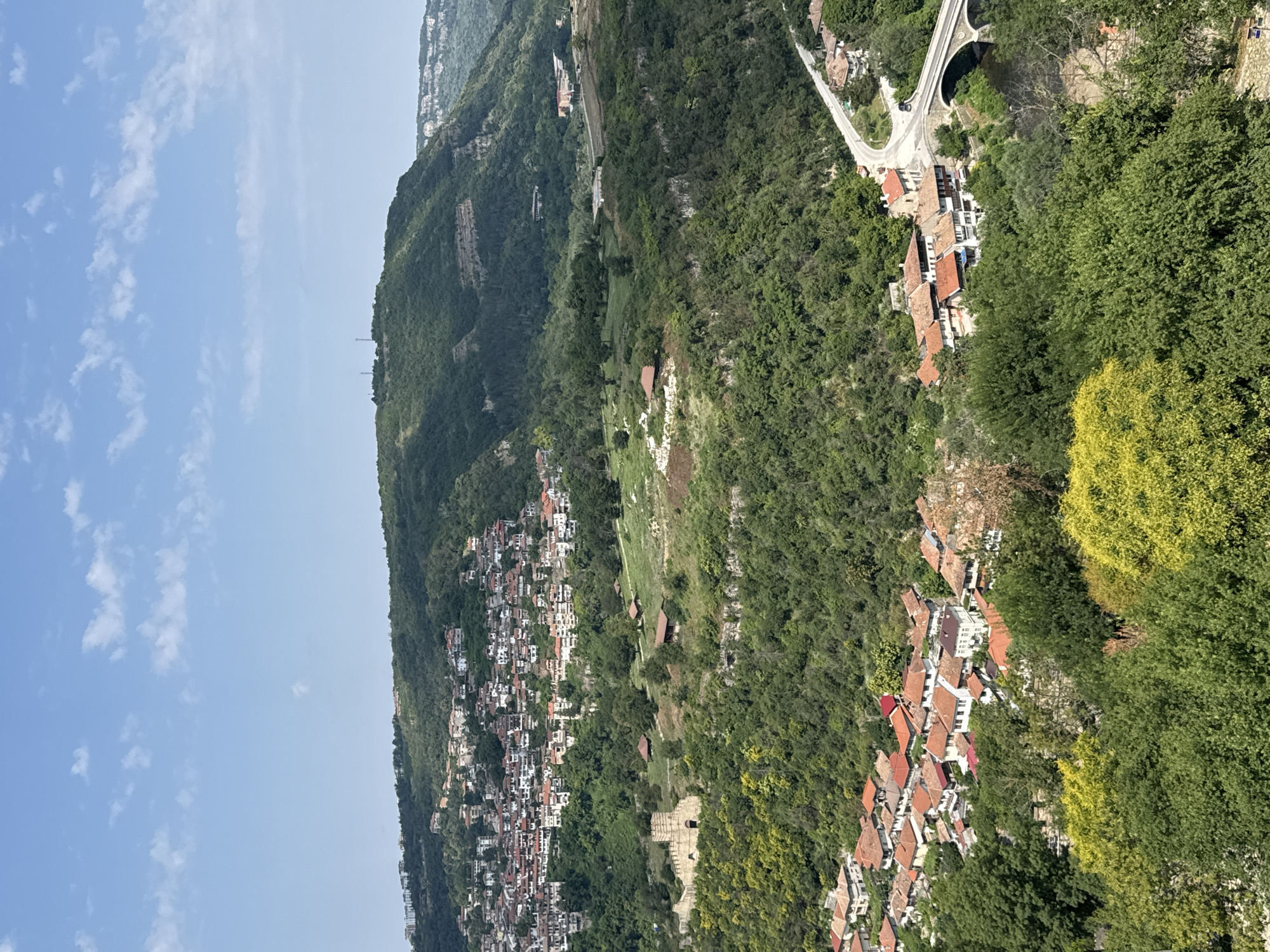 This is a high-angle, wide shot of Veliko Tarnovo, Bulgaria, on a clear day with scattered white clouds. The image captures a panoramic view of the city spread across rolling hills and nestled within a lush green valley. In the foreground, dense green foliage dominates, with patches of bright yellow-green trees adding color.
Moving further back, a cluster of traditional houses with red-tiled roofs is visible, densely packed together and built on the slopes of the hills. These houses are predominantly white with wooden accents. To the left, a portion of an ancient stone structure, likely ruins, is visible amidst the greenery.
The middle ground features more of the city, with houses cascading down the hillsides, interspersed with trees. A winding road is seen in the distance, leading towards a stone bridge that crosses a river. The bridge is elegant with multiple arches. The background reveals further hilly terrain, covered in dense green vegetation, with some scattered buildings and what appear to be telecommunication towers on the highest peak. The overall impression is of a historic city integrated with its natural surroundings.