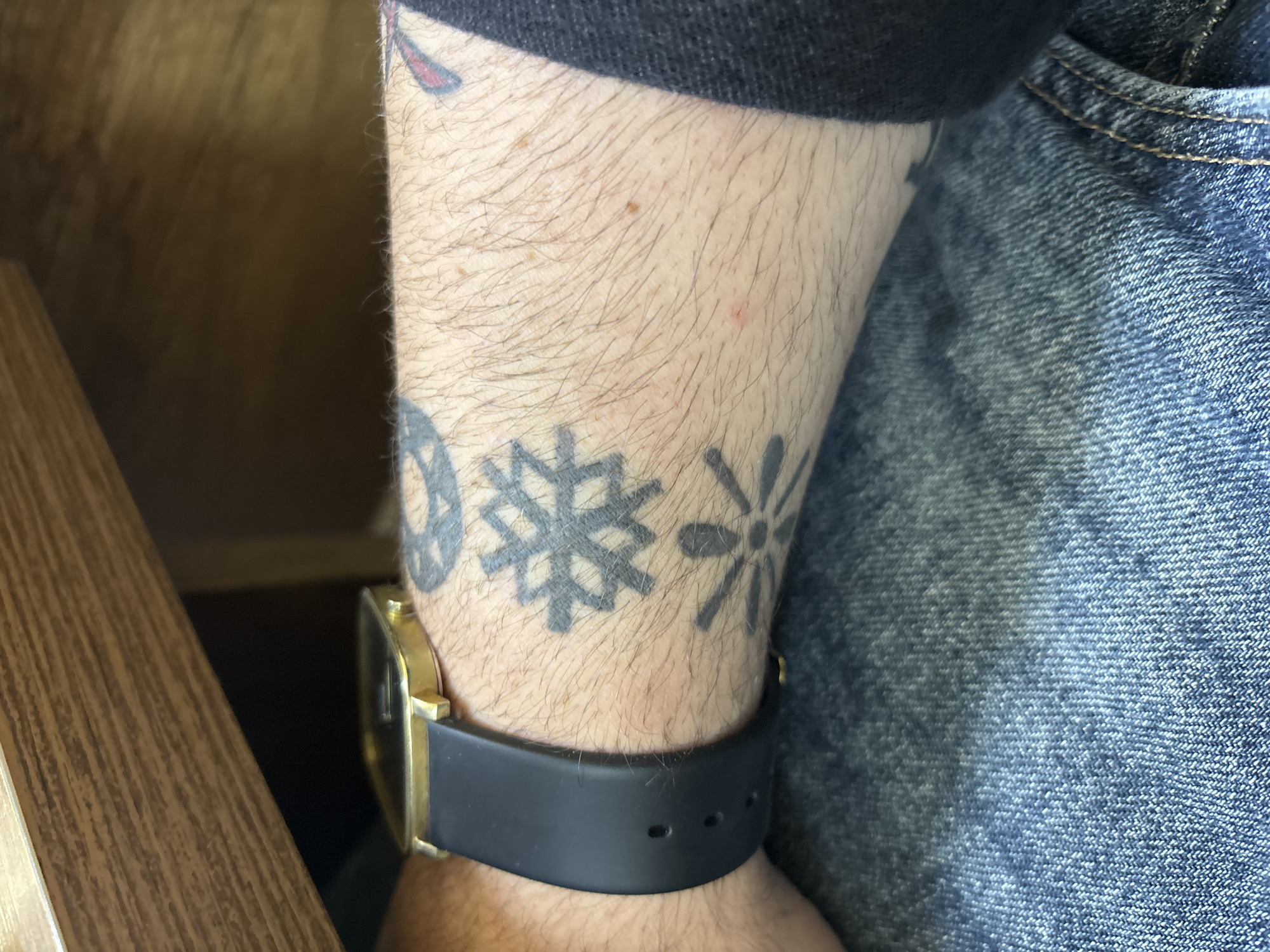 The image depicts a person's left forearm adorned with three circular, tattoo designs that are close together, resembling snowflakes or stylized flowers. The tattoos are black and appear slightly faded. The arm is hairy and has some small blemishes. A watch with a gold-colored case and a black band is worn on the wrist, positioned just below the tattoos. Part of a blue denim-clothed leg is visible on the right side of the image, possibly jeans. The background suggests an indoor setting with a portion of a wooden chair or table visible on the left. There is no indication or information that would identify this image as being taken in the city of Tbilisi, Georgia.