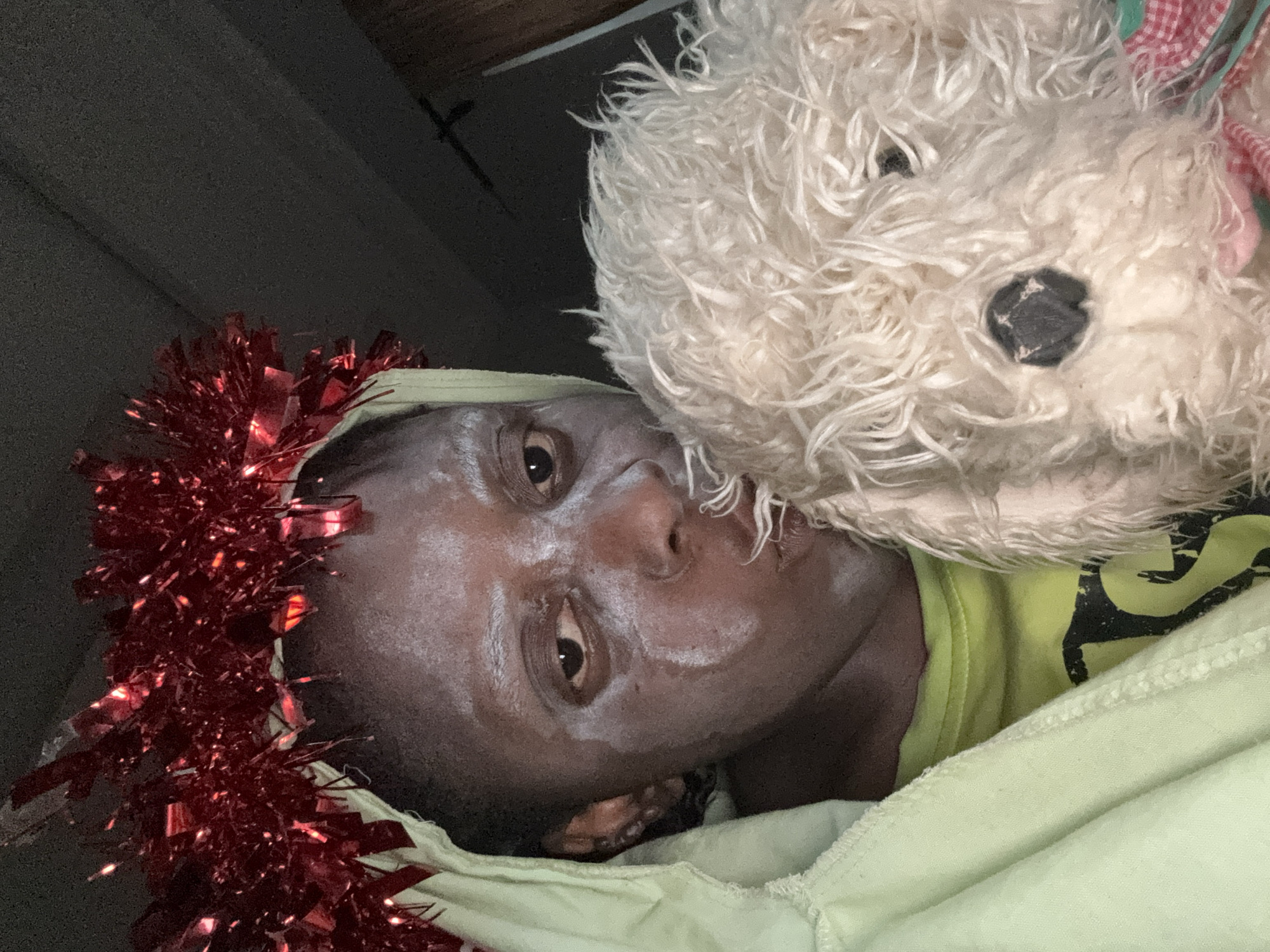 The image is a close-up selfie of a person lying down, face angled towards the camera. The person has dark skin, with white cream on their face. They are wearing a red tinsel garland around their head. A stuffed, white, furry dog toy is positioned next to their face. The person is wearing a light green garment, possibly a hoodie or blanket. The environment appears to be indoors, with dark ceiling beams visible in the background. The location of Sabon Tasha, Nigeria is not explicitly evident in the image. The image contains no actions or interactions between people.