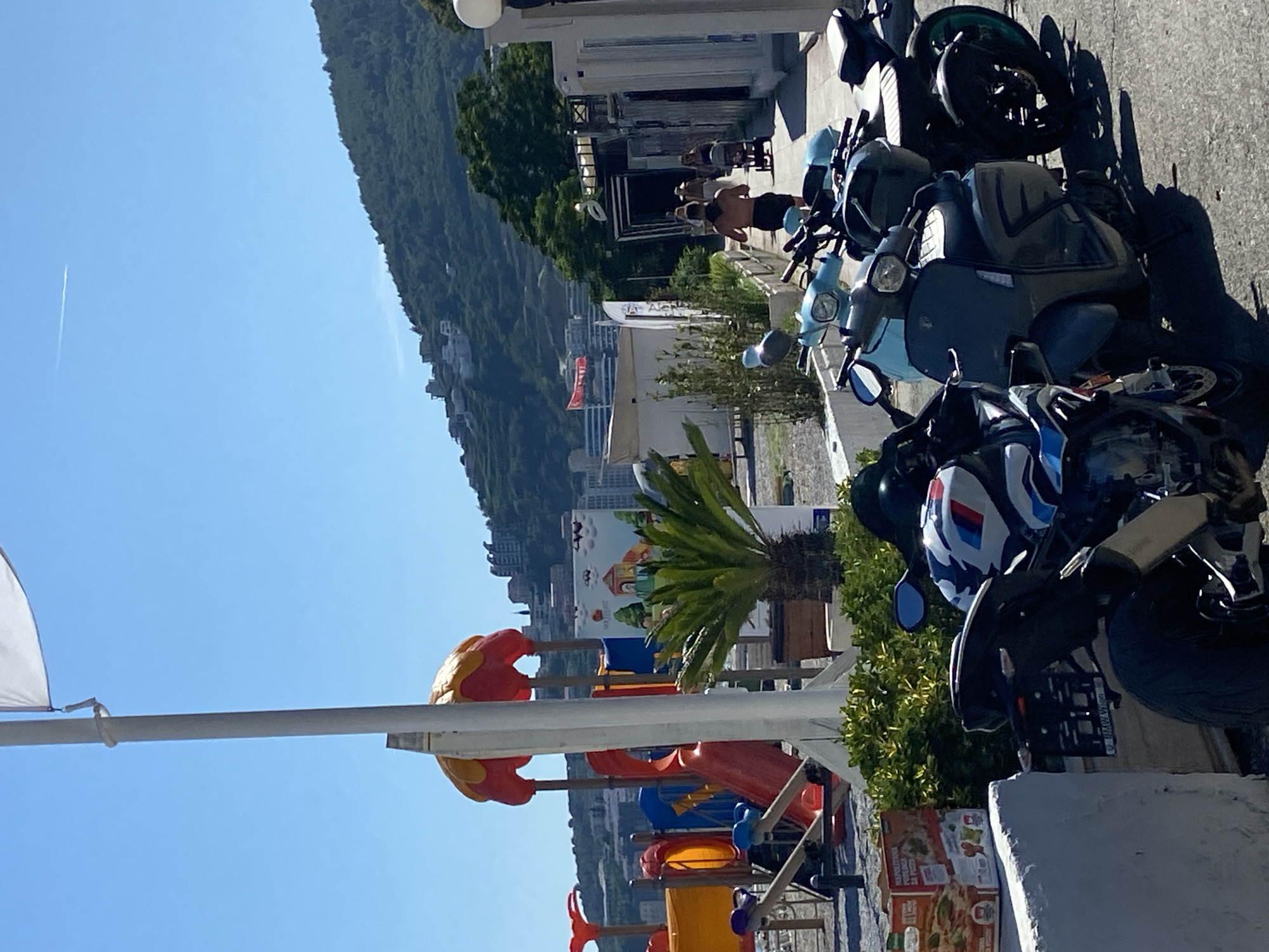 The image depicts a sunny day scene, likely in Budva, Montenegro, as suggested by the context. In the foreground, several parked motorcycles and scooters are visible on a paved surface. One motorcycle, in particular, has a distinctive white, blue, and red livery. Behind the vehicles, a playground with colorful slides and structures is set up, suggesting a public recreational area. Further in the background, a lush green hillside is dotted with buildings, and a bright blue sky stretches above, with a faint contrail from an aircraft. Several people are seen walking in the distance, some pushing strollers, along a walkway bordered by greenery and architectural elements. A red flag is visible on a building on the hillside. The overall impression is a vibrant, outdoor setting with recreational and residential elements.