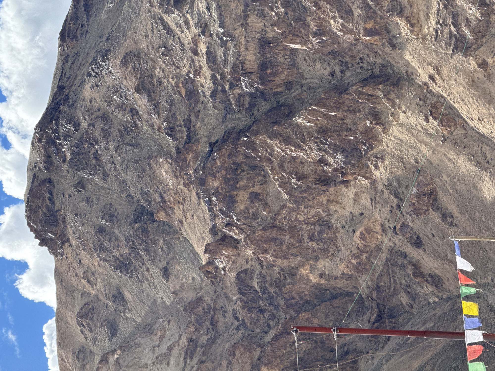 The image depicts a rugged mountain landscape with a blue sky and scattered white clouds. In the foreground, a red pole stands vertically, with wires stretching horizontally across the frame. Below the pole, a string of colorful prayer flags is visible, including white, red, blue, yellow, and green flags. The mountain itself is characterized by its dry, rocky terrain, with shades of brown, tan, and gray dominating its surface. The texture of the mountain is rough, with visible rocks and scree. The lighting suggests it is daytime, with sunlight casting shadows and highlighting the contours of the mountainside. The prayer flags and utility pole indicate human presence in this otherwise wild environment, hinting at a settlement or a path in the vicinity of Koyul, India.